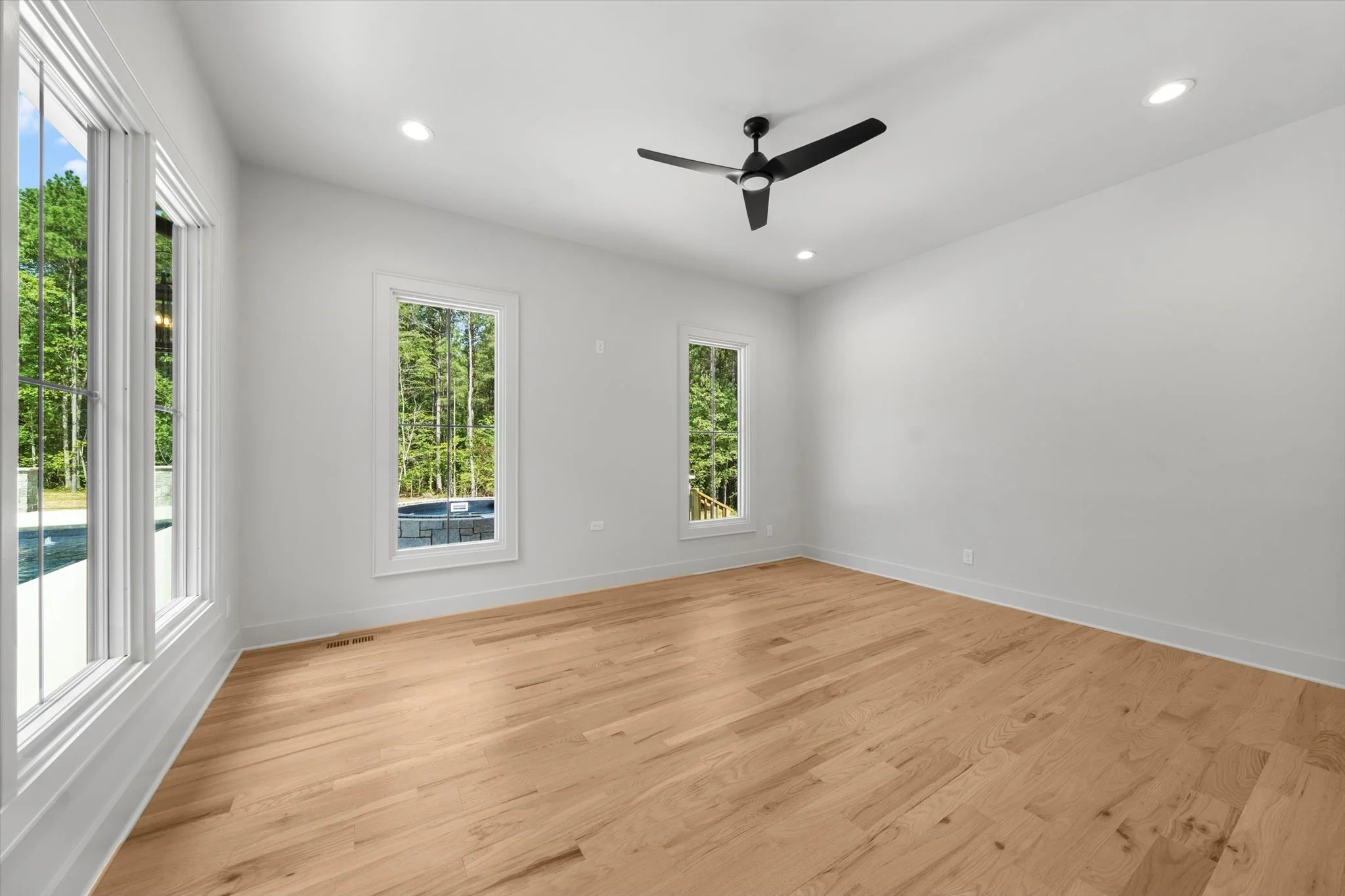 Empty room with light wood floors, white walls, three large windows, a ceiling fan, and recessed lighting, with a view of greenery outside.