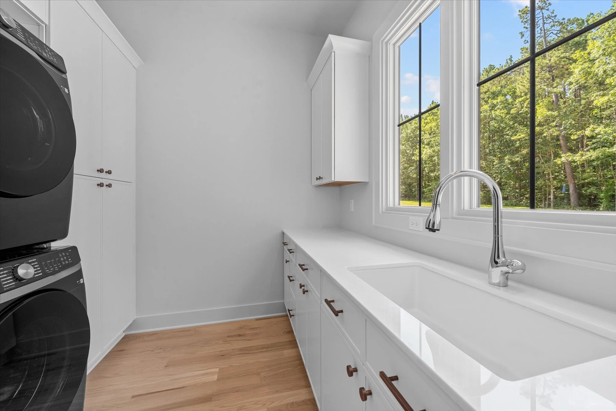 Laundry room with white cabinets, a large white countertop with a sink, a chrome faucet, a large window overlooking a green wooded area, and a stacked washer and dryer in black.
