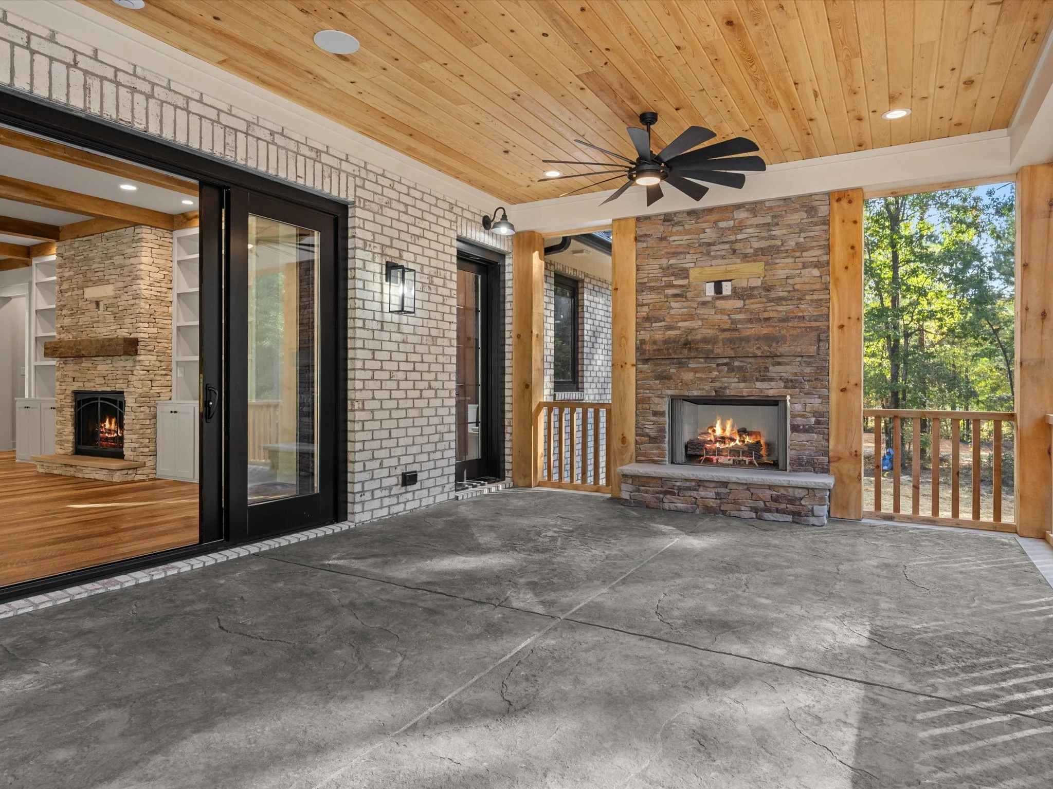 Covered patio with concrete flooring, brick and stone walls, two fireplaces, ceiling fan, sliding glass doors, and large windows surrounded by wooden beams and a scenic view of trees outside.