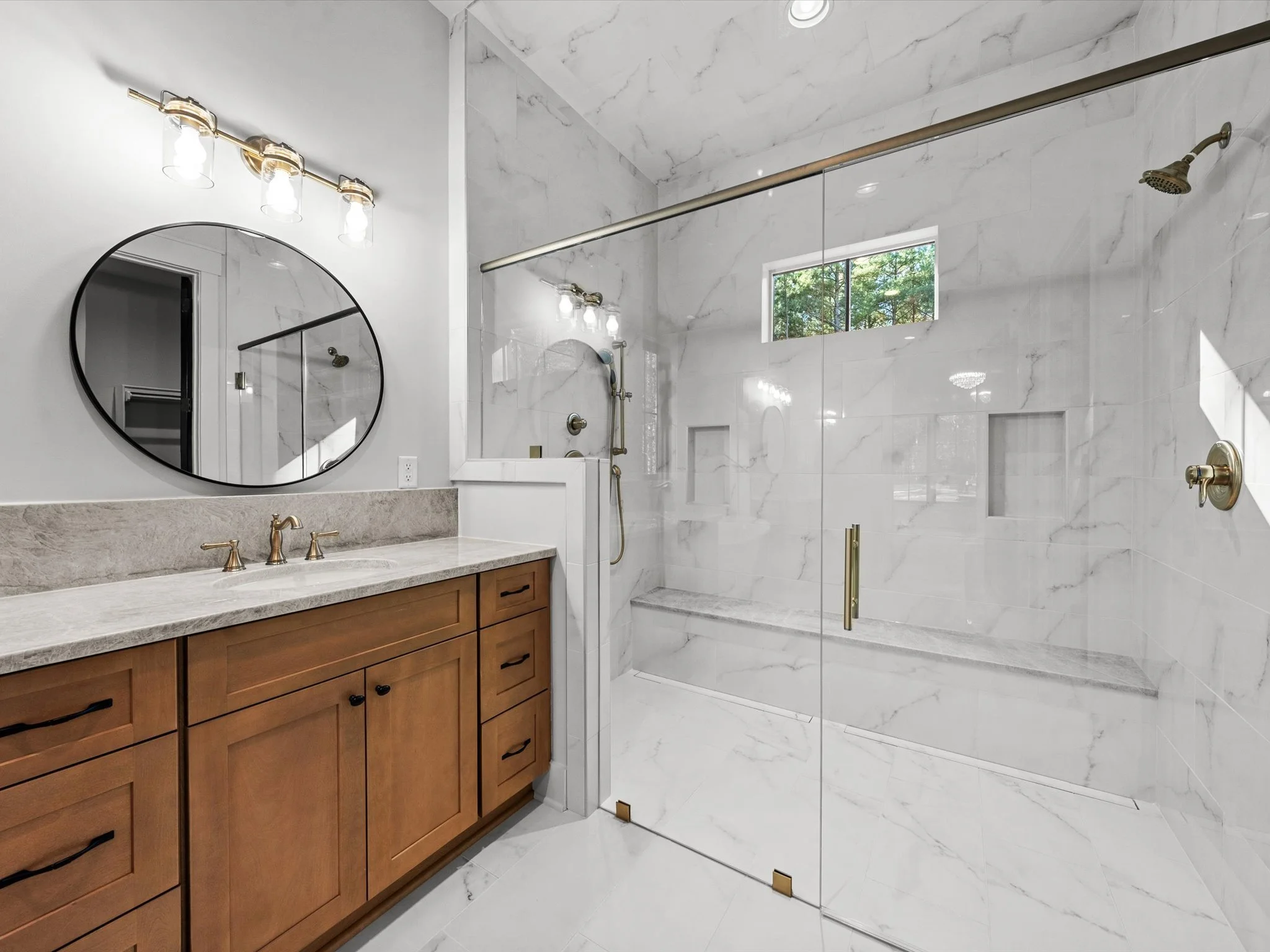 A modern bathroom featuring a wooden vanity with a marble countertop, a round mirror, and a spacious glass-enclosed shower with marble tiles, a bench, and built-in niches.