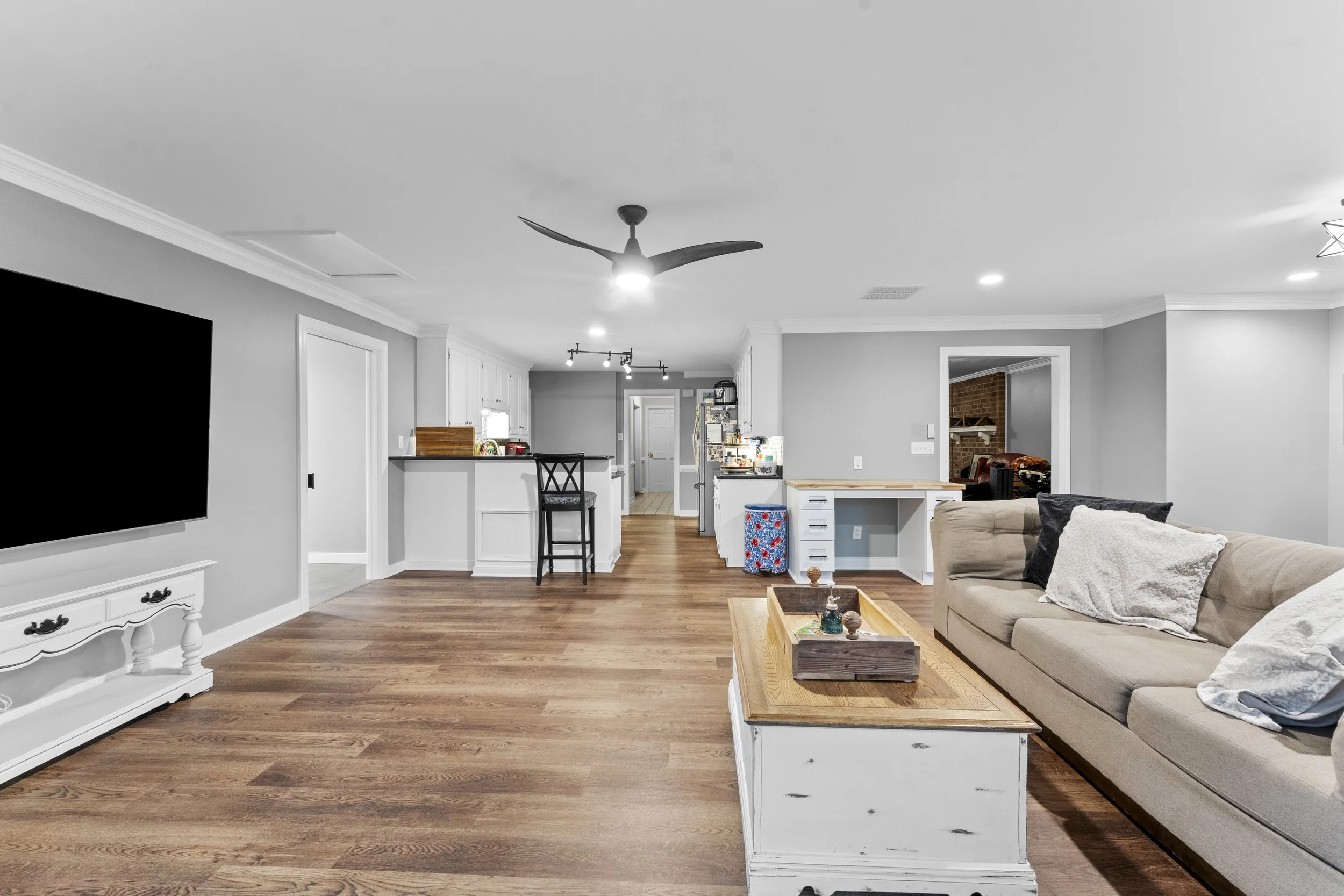 An open-concept living room and kitchen with hardwood floors, a beige sofa with pillows, a white and wood coffee table, a wall-mounted flat-screen TV, a ceiling fan, and kitchen appliances in the background.