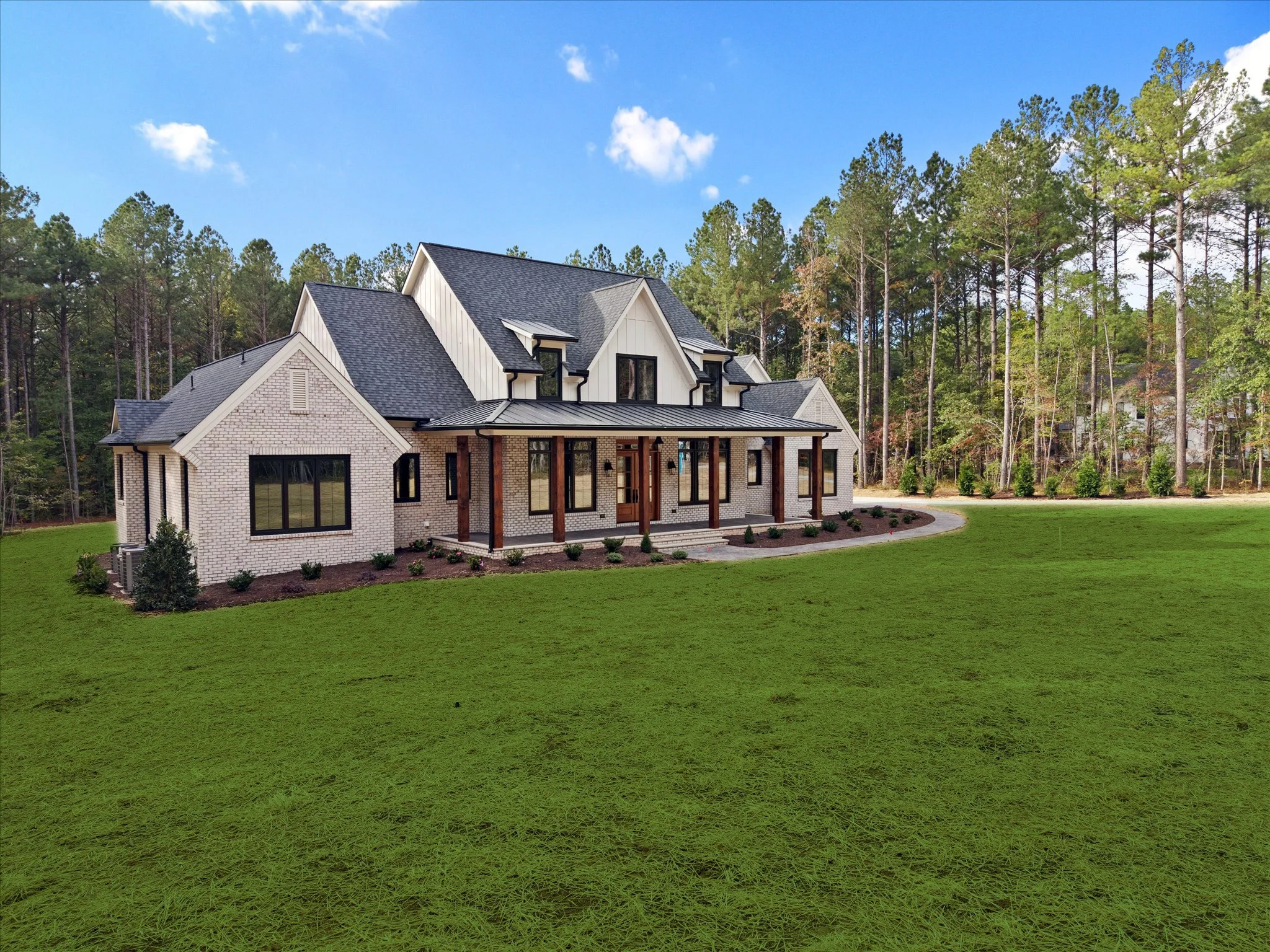 A modern white brick house with a black shingle roof, multiple gables, and large windows, surrounded by a lush green lawn and a wooded area with tall trees in the background.