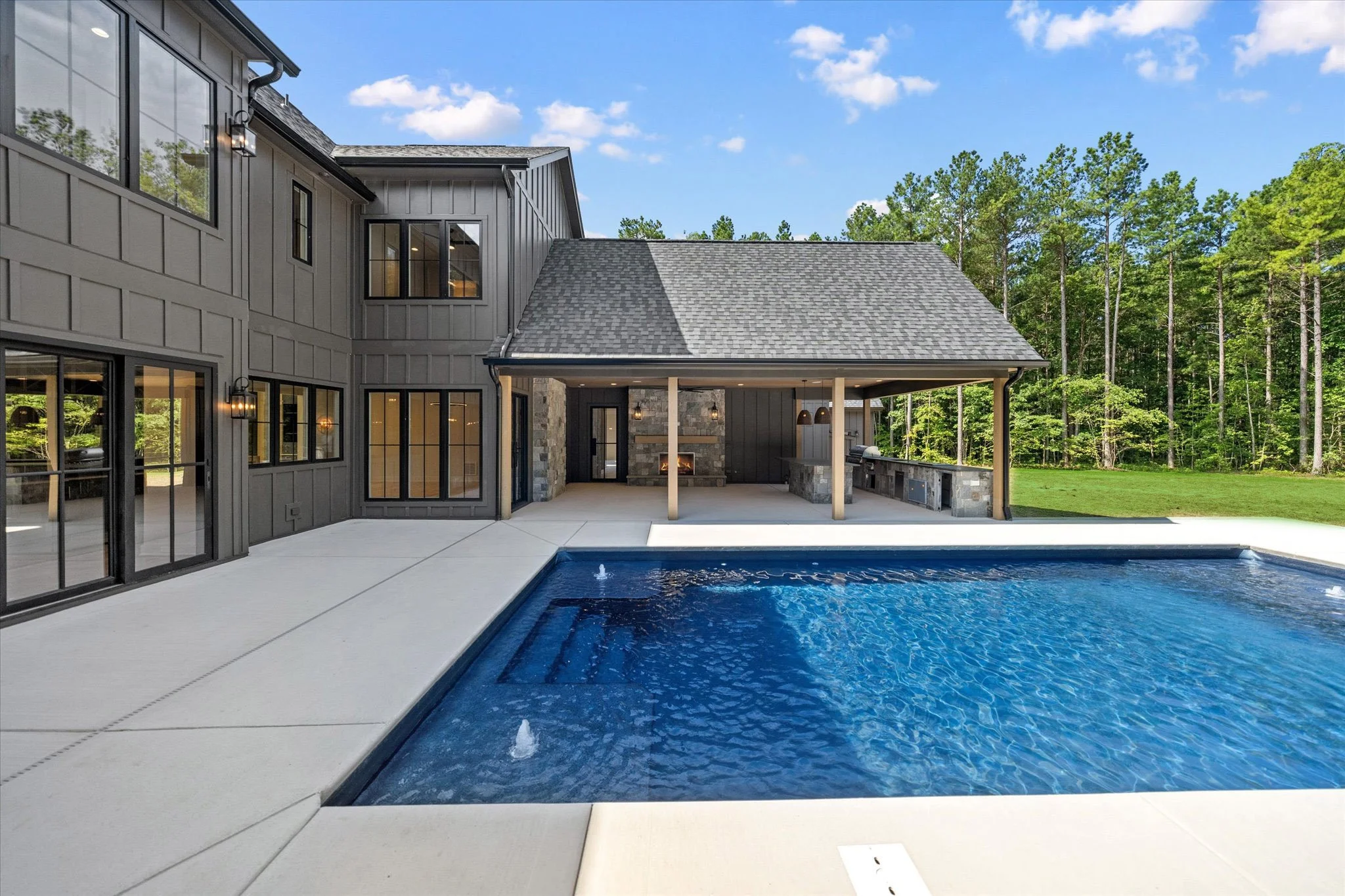 A backyard with a modern house featuring large windows, an outdoor covered patio with a fireplace, and a swimming pool with water features, surrounded by green trees.