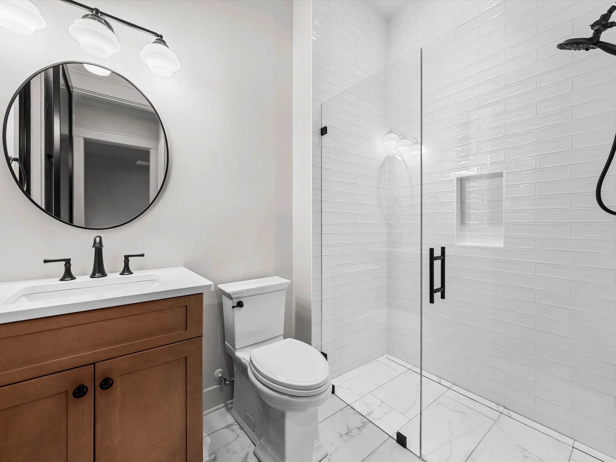 Bathroom with a wooden vanity, white countertop, black fixtures, a porcelain toilet, a large round mirror, white tiled shower with a glass door, black handle, and showerhead, marble tile flooring.
