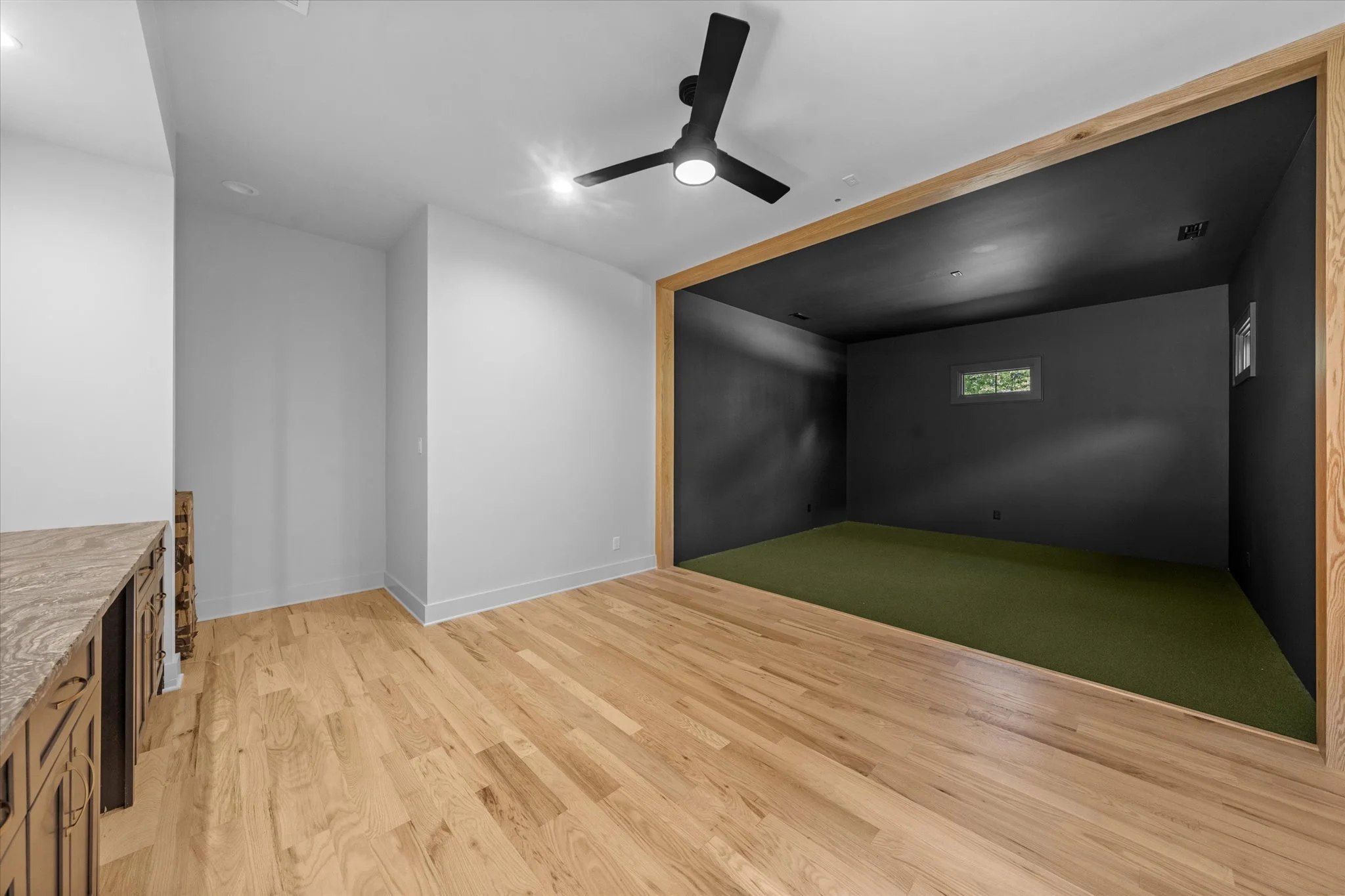 A spacious room with light-colored wooden floors, a white wall, a ceiling fan, and a dark-colored alcove with a green carpet and a small window.