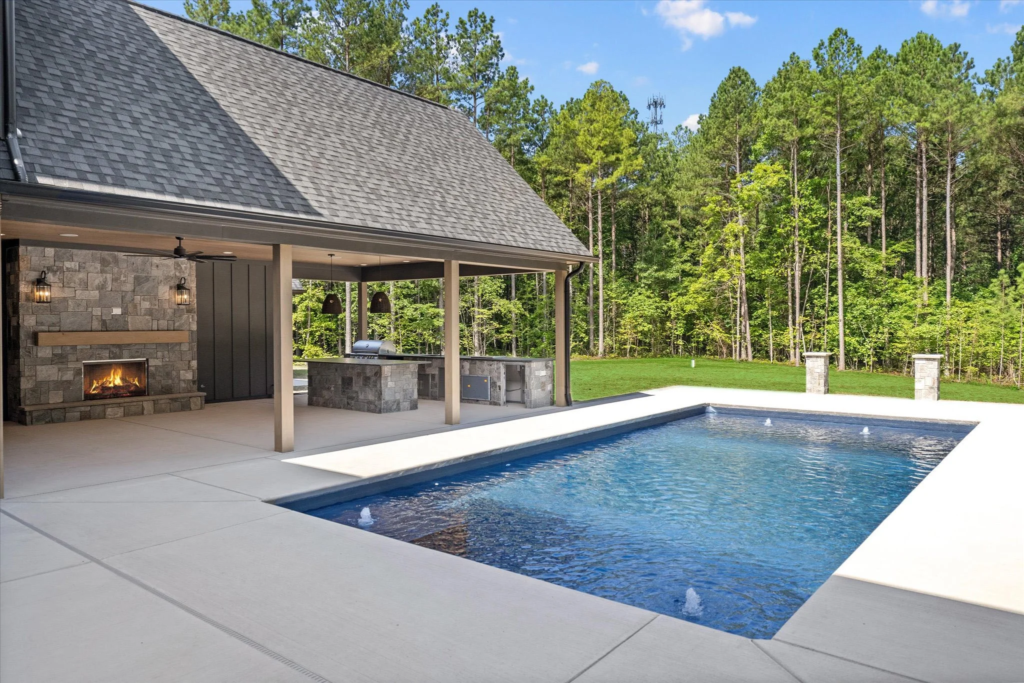 Modern backyard with a swimming pool, outdoor fireplace, covered patio with a built-in grill and seating area, surrounded by green trees under a blue sky.