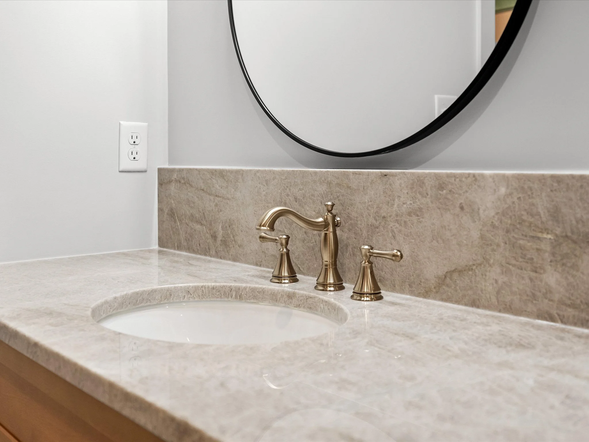 Bathroom sink with a beige marble countertop, brass faucet, and a round mirror on a light gray wall.