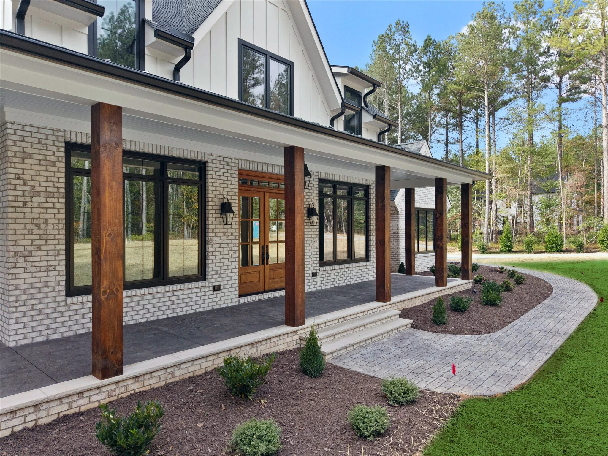 Front porch of a modern house with brick and wood exterior, black framed windows, and a wooden door, surrounded by landscaped garden and pathway.