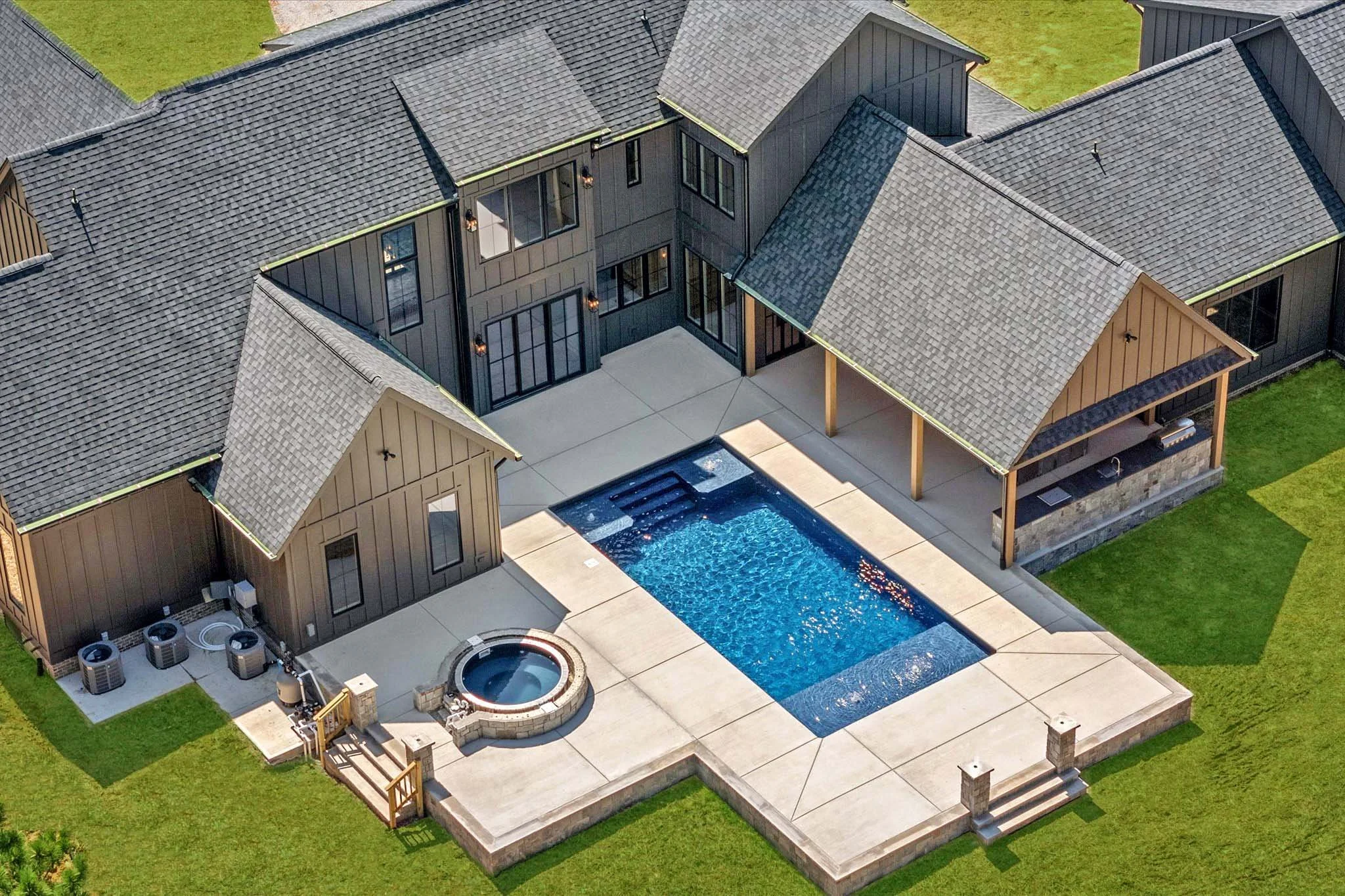 Aerial view of a modern house with a backyard featuring a rectangular swimming pool, a small hot tub, and a covered patio area, surrounded by green lawn.