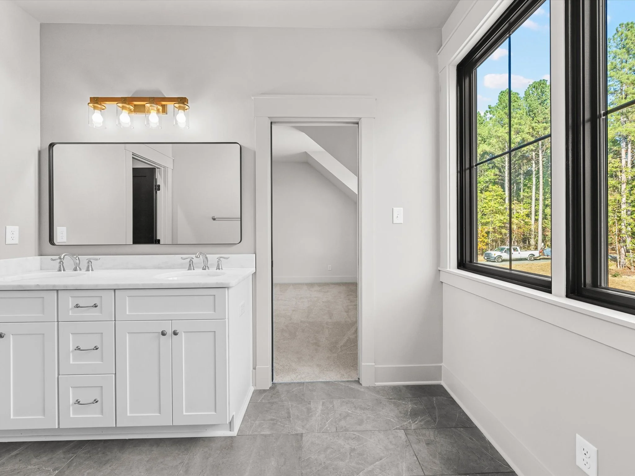 Bathroom interior with white vanity, double sinks, large mirror, and four-bulb light fixture. Large window shows a view of trees and parked cars.