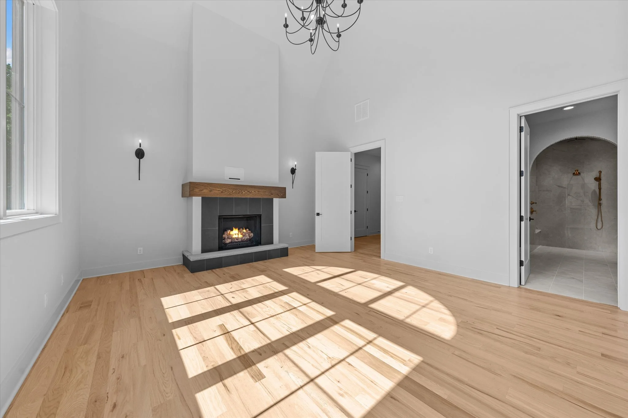 Empty living room with hardwood floors, a fireplace with a wooden mantel, wall-mounted lights, large window casting sunlight on the floor, and a doorway leading to a bathroom with a shower.