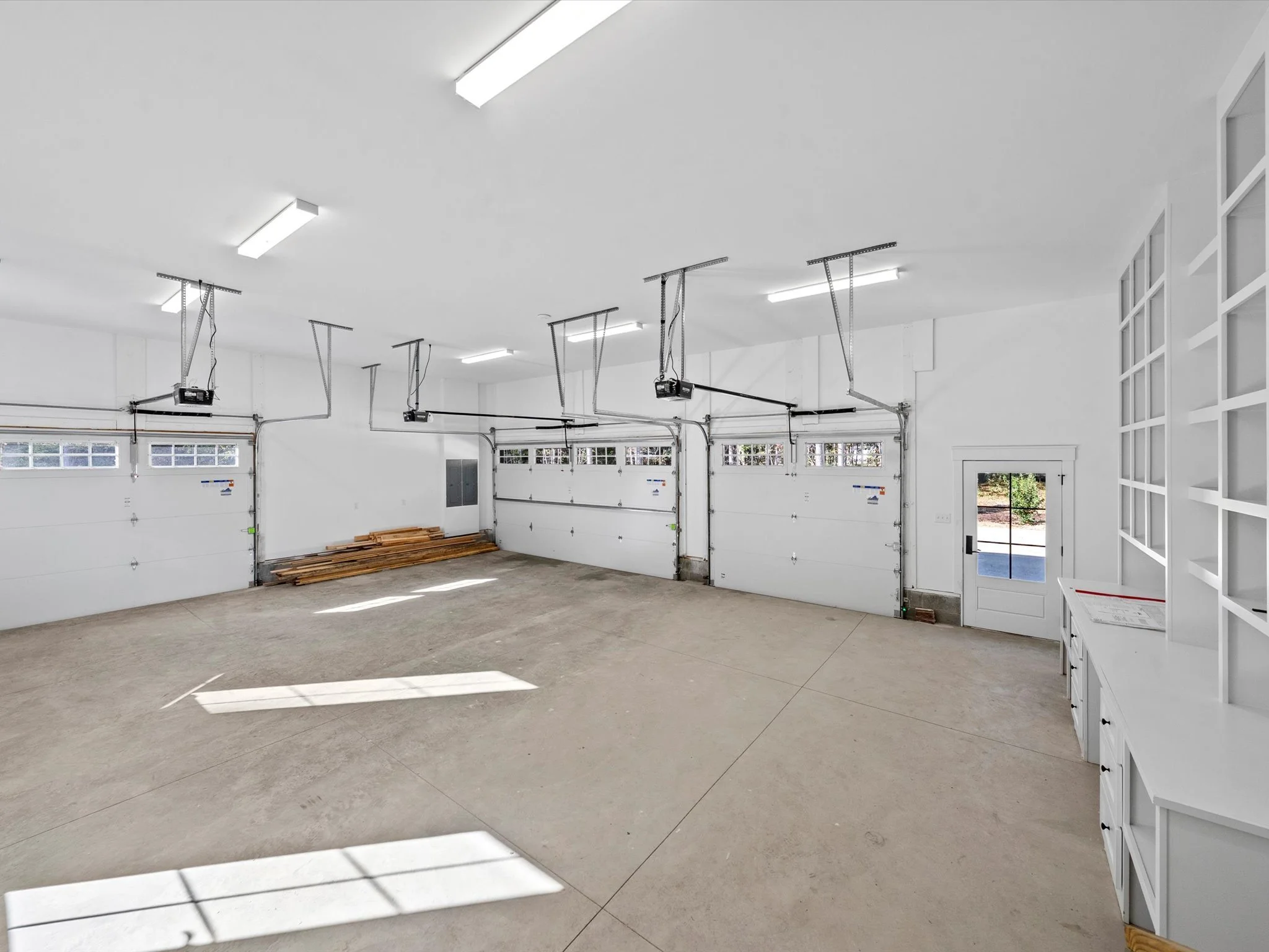 Empty clean garage with three white garage doors, overhead garage door openers, a small side door, and some stacked wooden planks. Bright lighting and sunlight streaming through the windows.