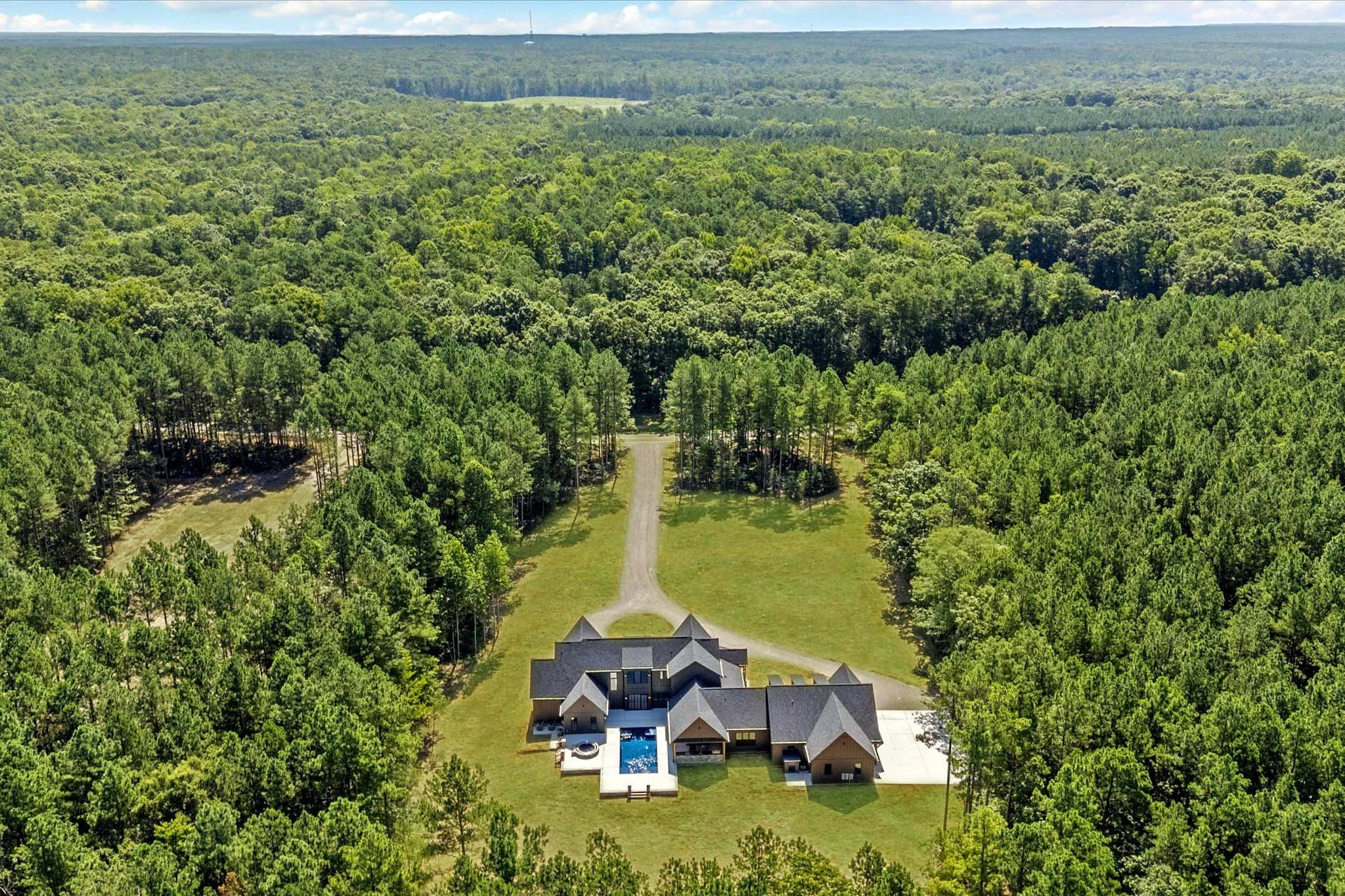 A large house with a swimming pool located in a spacious grassy and tree-filled area, set in a heavily wooded landscape with dense forest surrounding it.