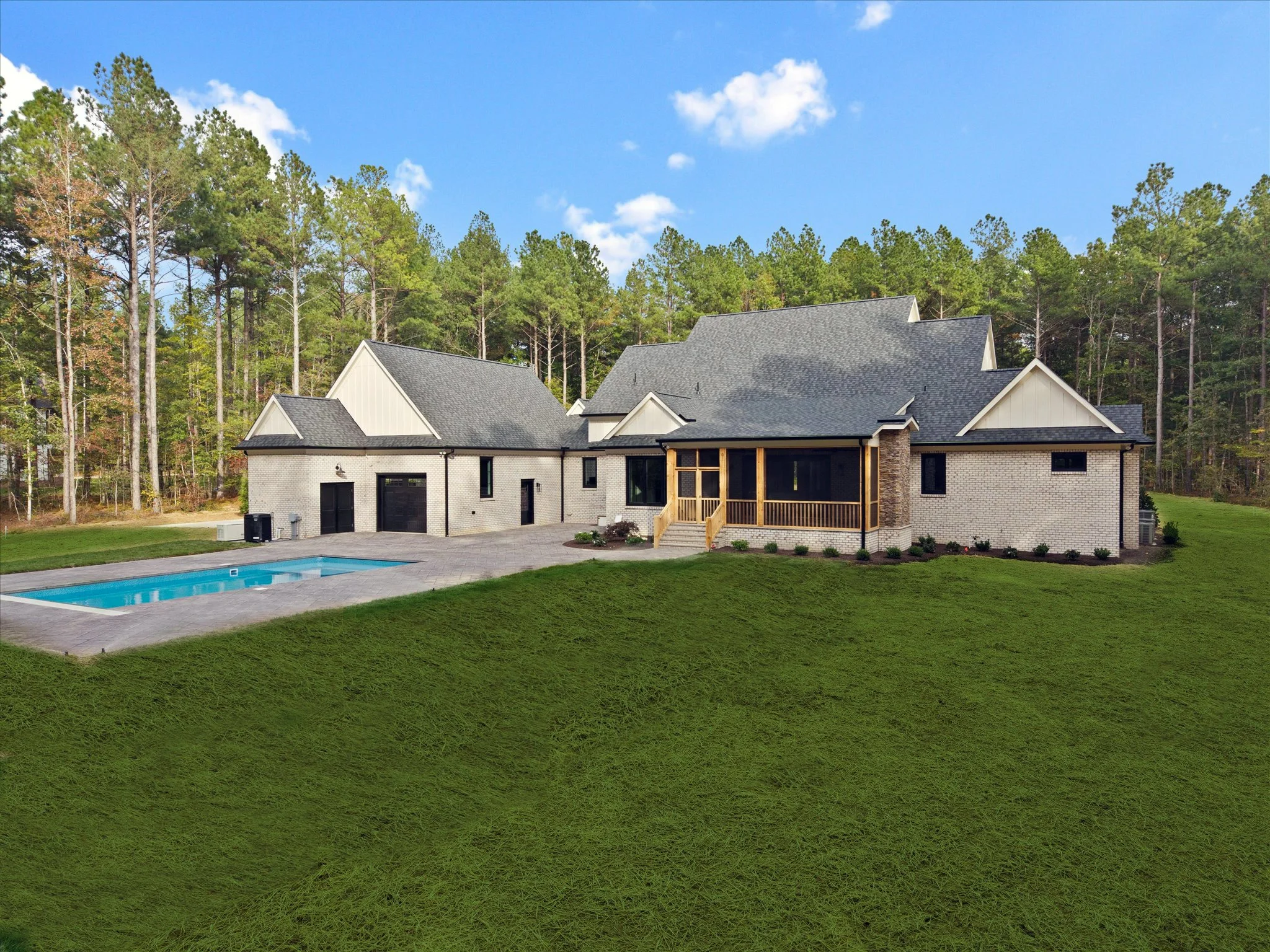 A modern house with white brick exterior, multiple gable roofs, a porch with wooden pillars, and a swimming pool in the backyard surrounded by green grass and trees under a blue sky.