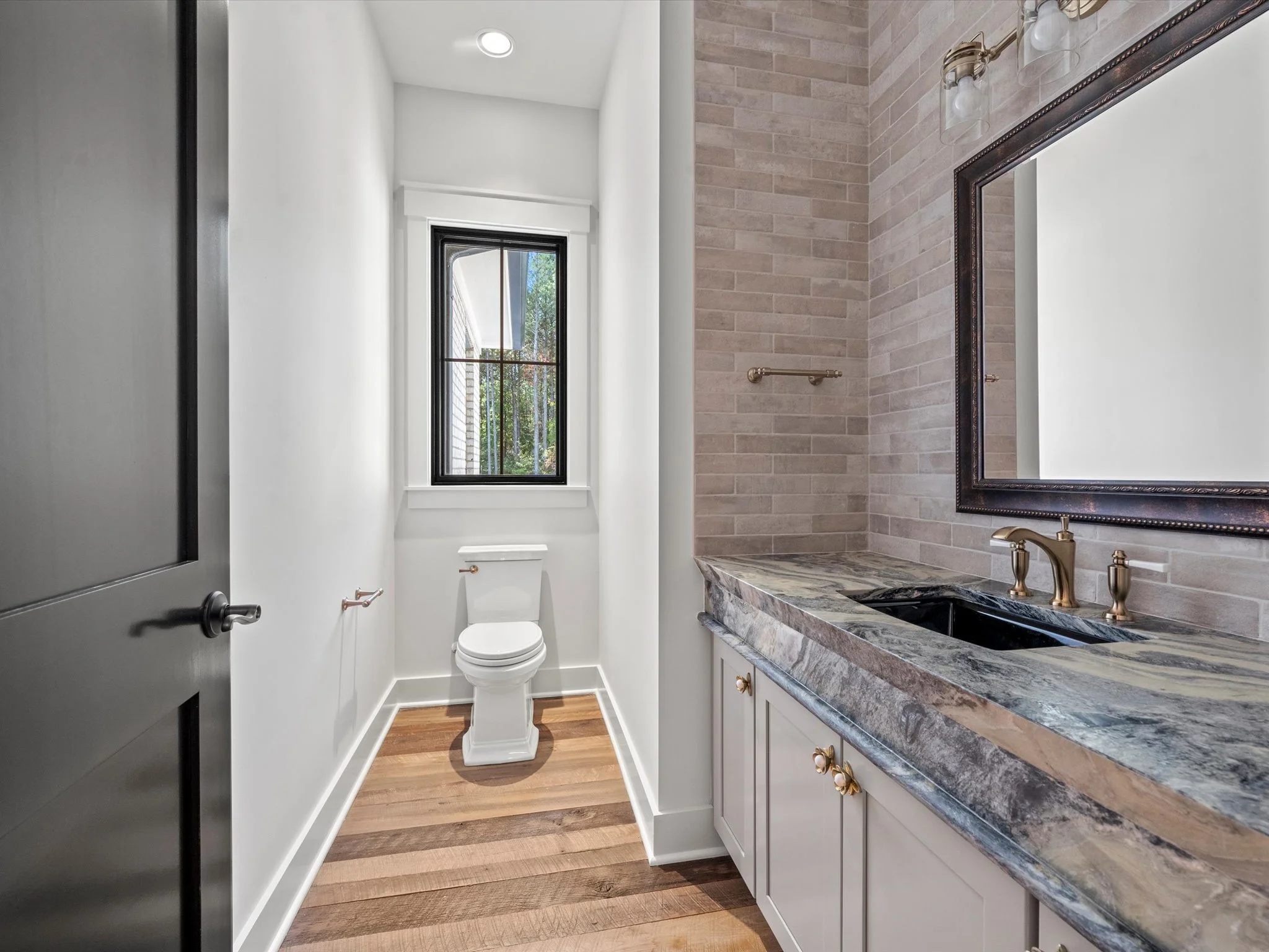 Modern bathroom with a white toilet, a marble countertop sink with gold fixtures, a large mirror with a dark frame, and a window with black trim overlooking greenery.