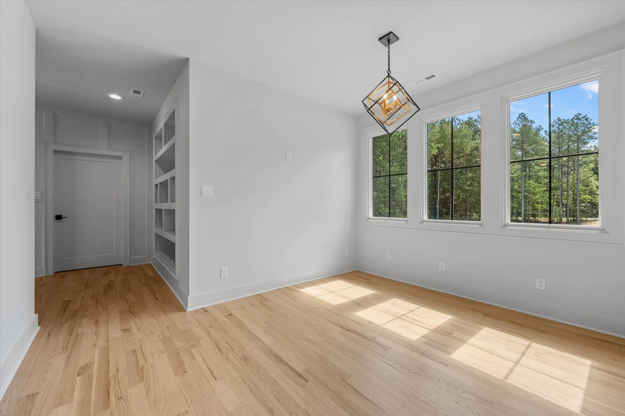 Empty room with three large windows showing trees outside, hardwood floors, white walls, and a geometric pendant light fixture hanging from the ceiling.
