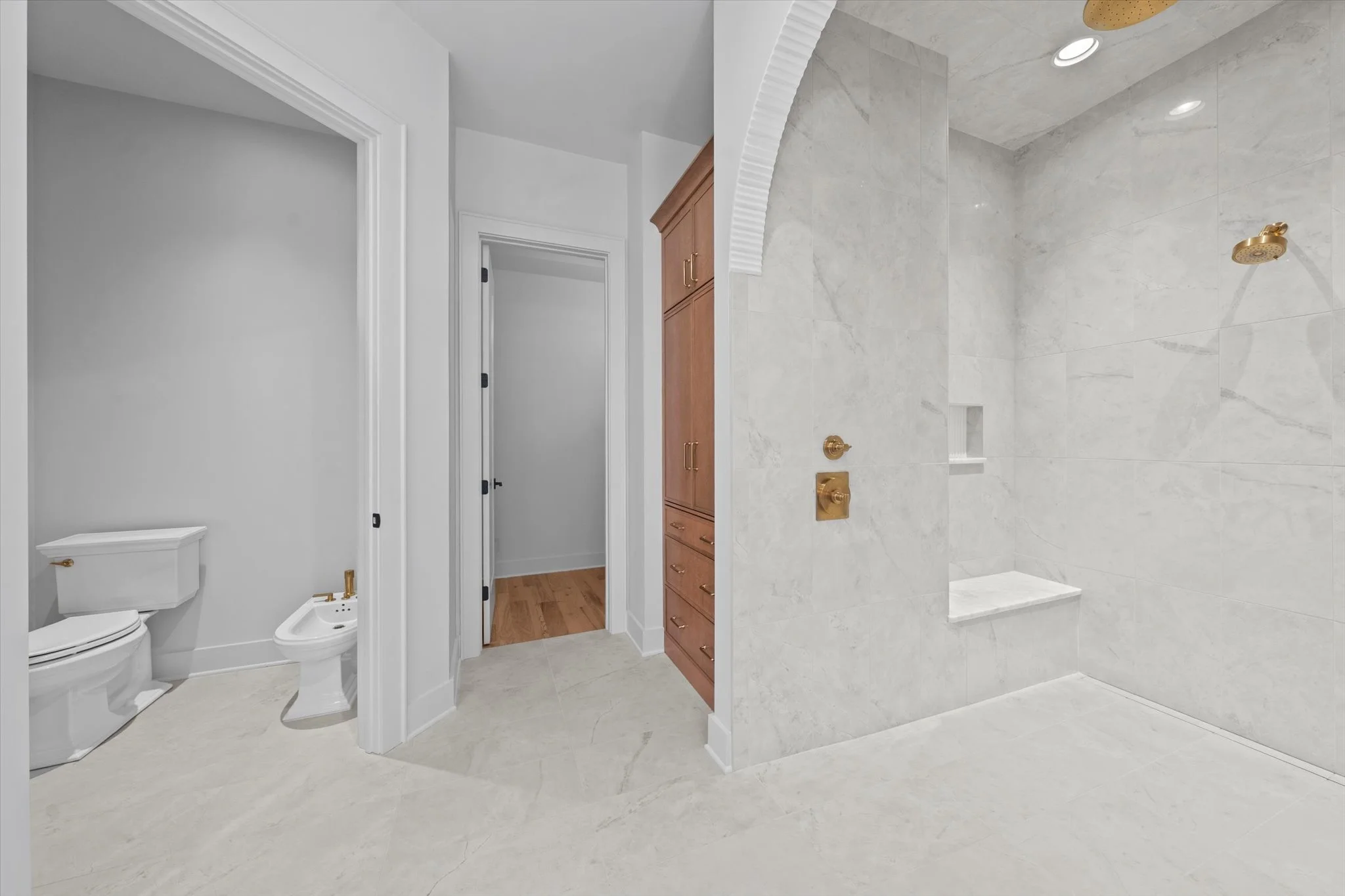 A clean, modern bathroom with white walls, a separate toilet and bidet, an open shower with gold fixtures, marble tiles, and wooden cabinets.