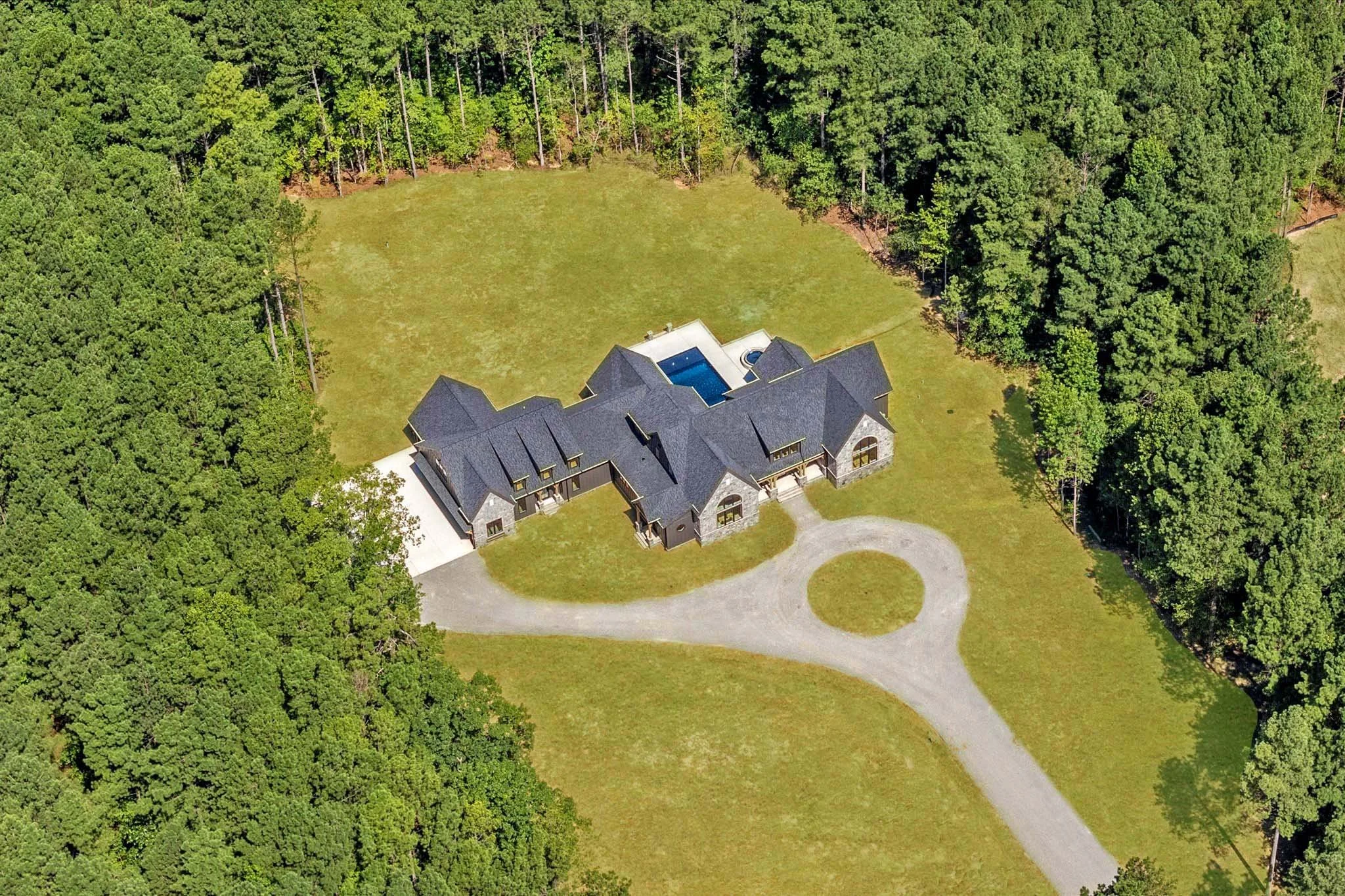 A large house with a dark roof surrounded by a spacious yard and dense trees, featuring a swimming pool in the backyard.