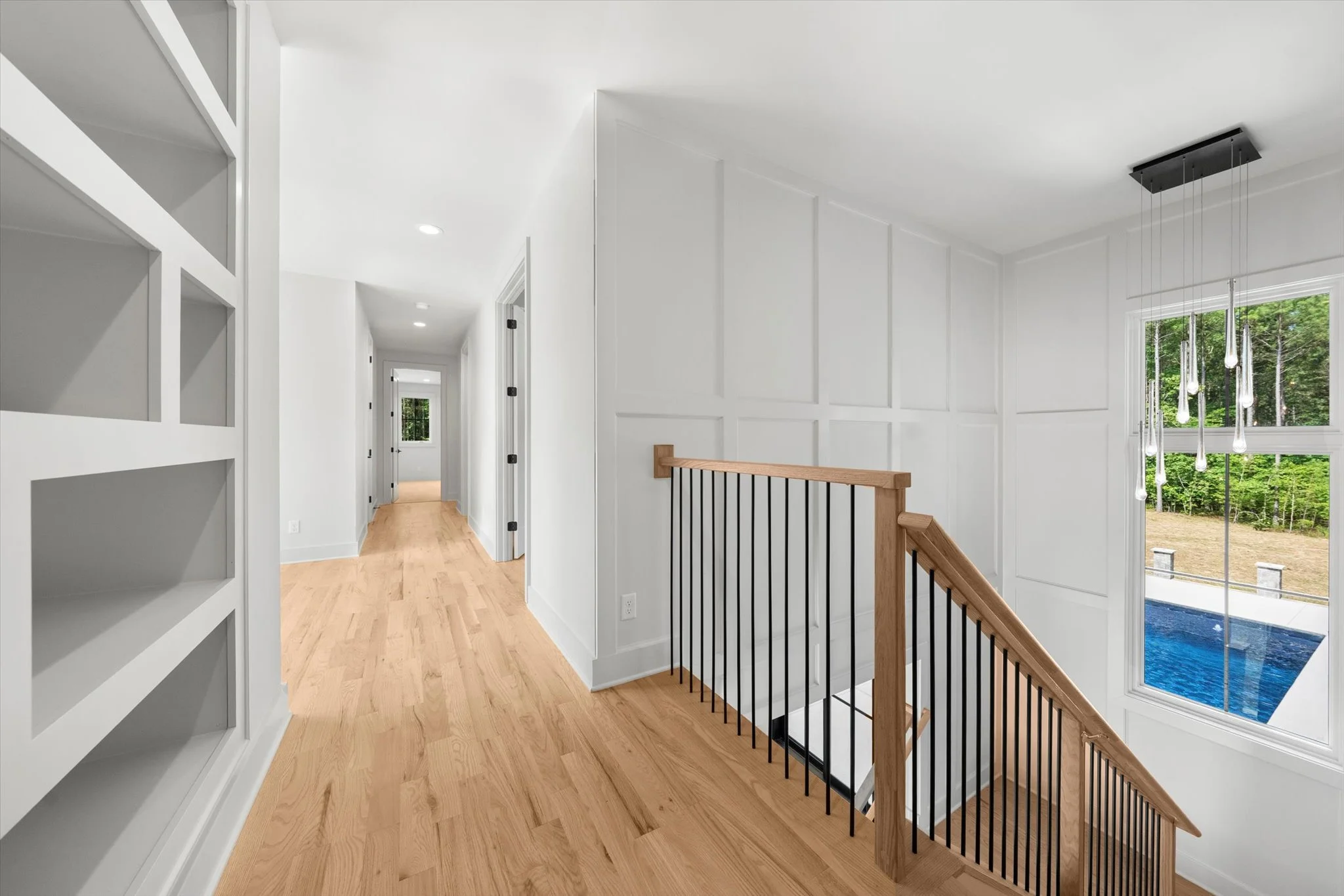 Contemporary home hallway with white walls, wooden floors, and a staircase with wooden handrail and black balusters. A large window reveals a backyard with a blue pool and green trees.