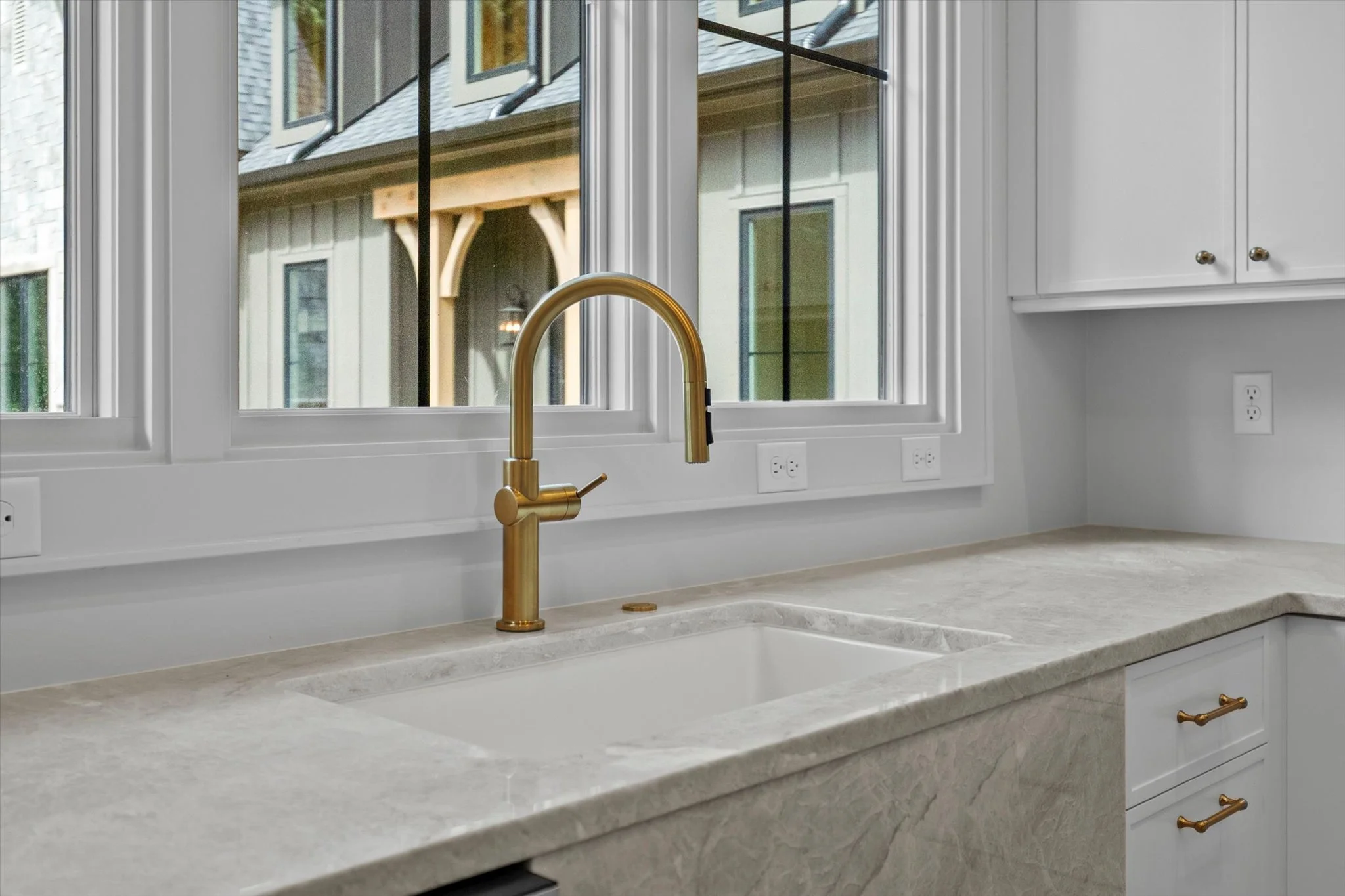 Kitchen sink with a gold faucet in front of a window showing neighboring houses.