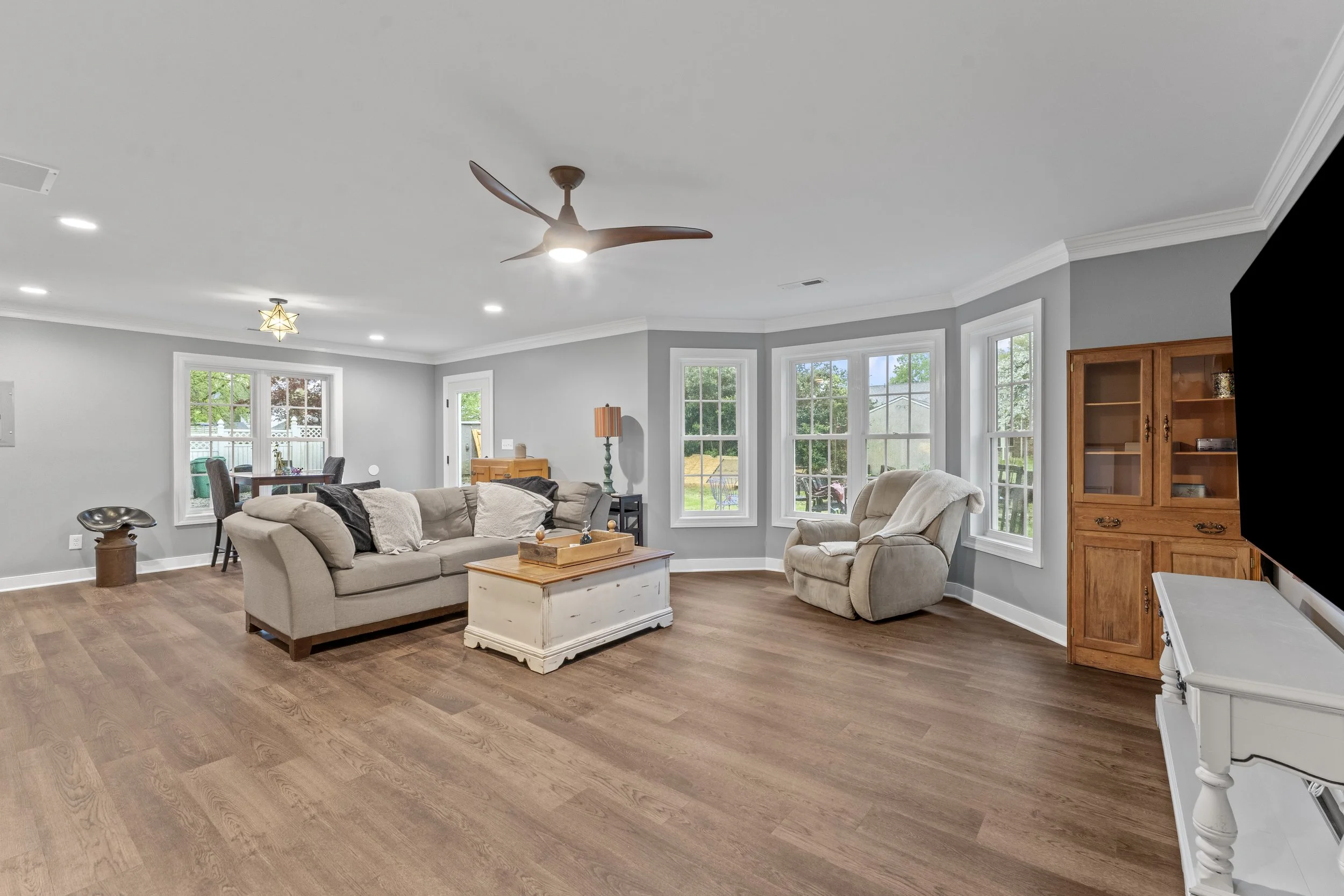 Living room with gray walls, hardwood floors, beige sofa and recliner, white coffee table, wooden cabinet, and large windows with a view of the backyard.