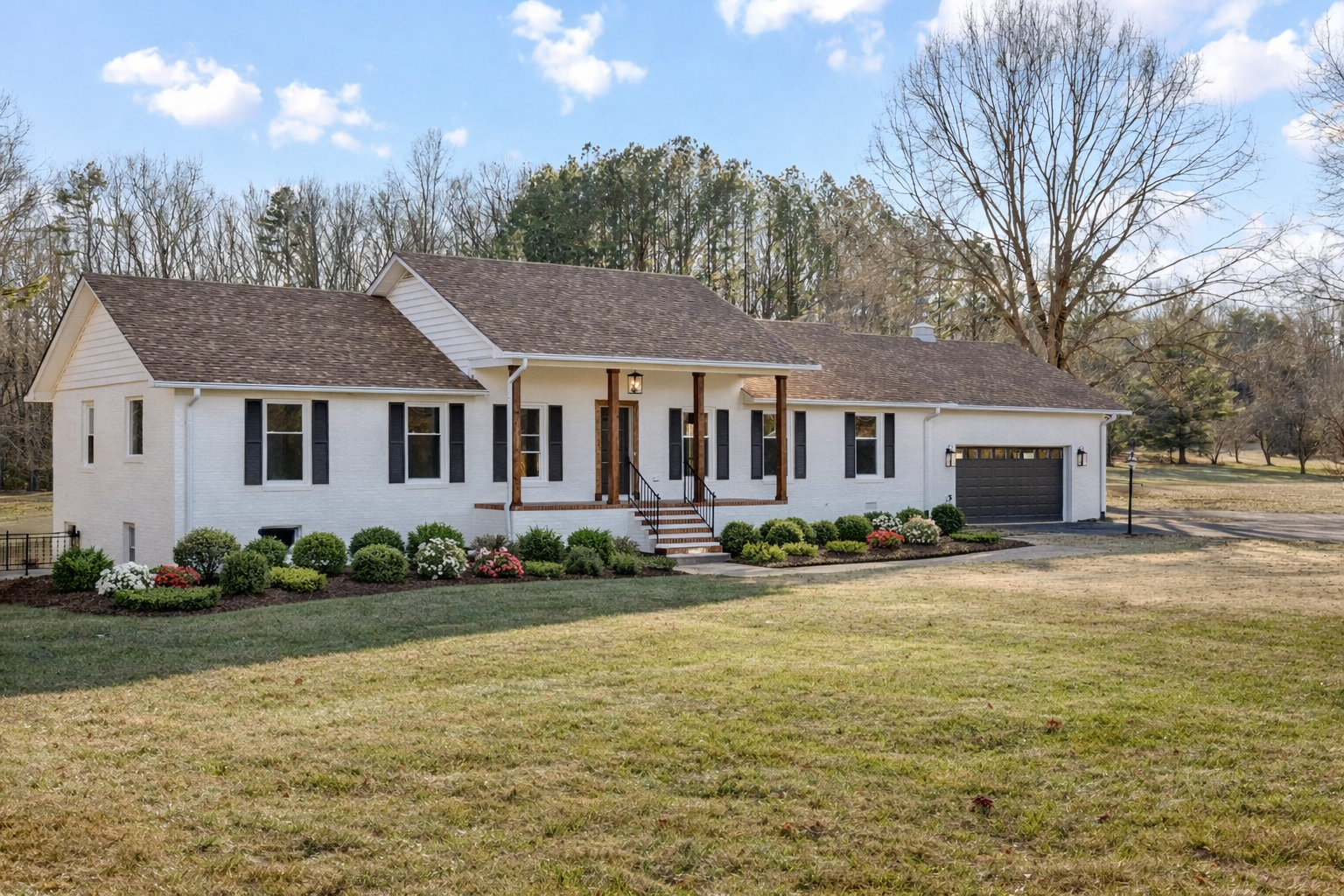 A white house with black shutters, a brown shingled roof, and a black garage door, surrounded by well-maintained flower beds and a large grassy lawn, with leafless trees in the background under a partly cloudy sky.