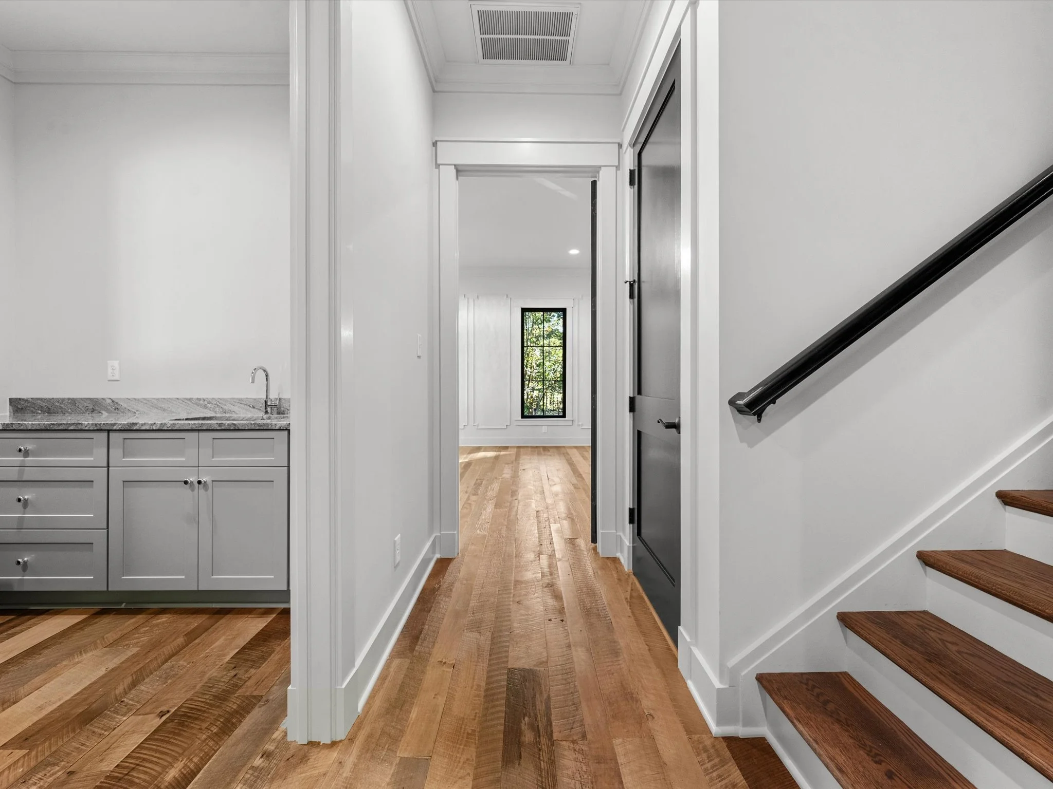Interior view of a modern home with hardwood floors, white walls, a staircase with wooden steps and a black handrail, a laundry area with gray cabinets and a granite countertop, a door on the right, and a room with a window at the end of the hallway.