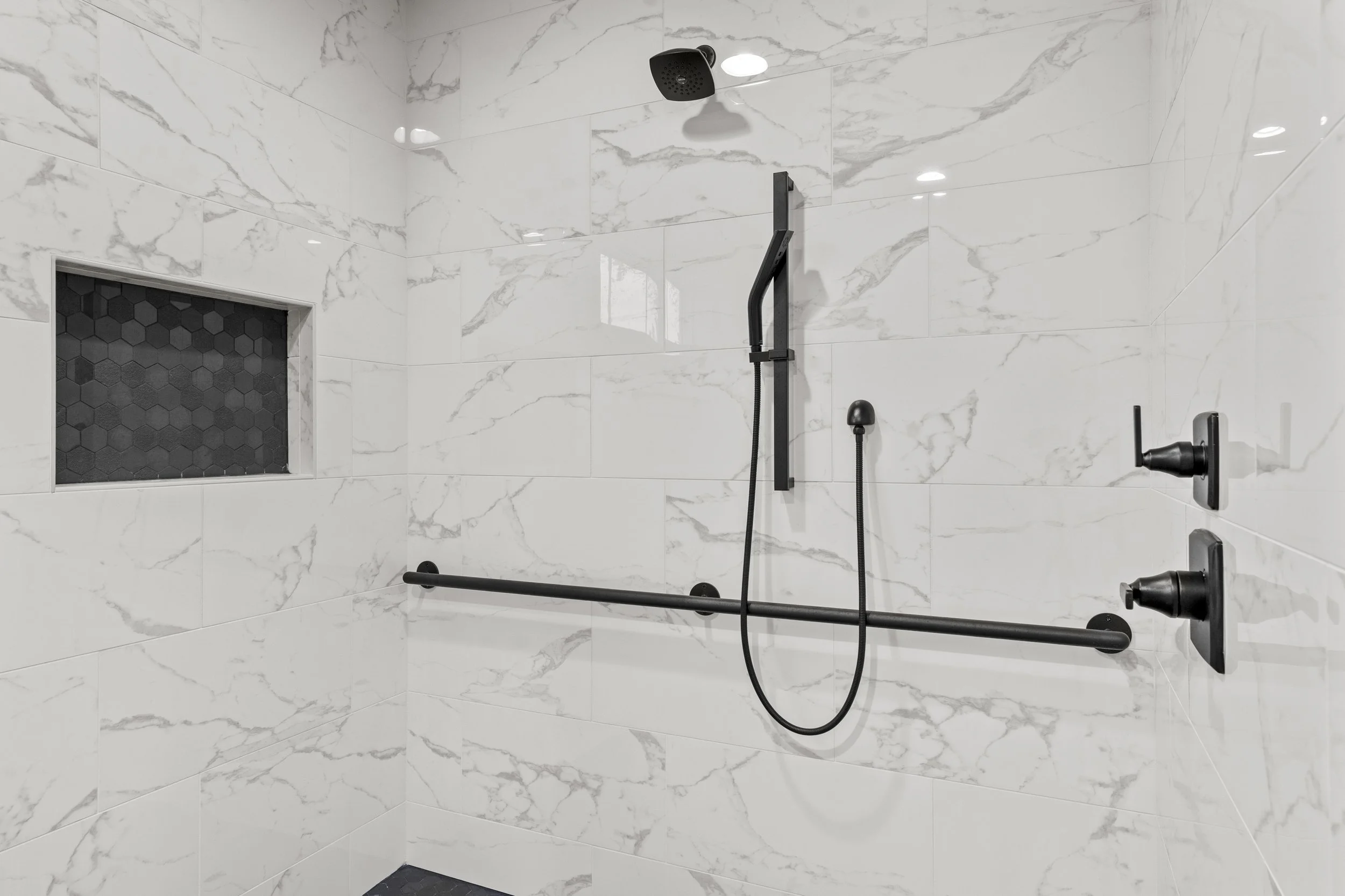 Accessible shower with white marble tiles, black grab bars, black shower head, and a built-in niche with black hexagonal tiles