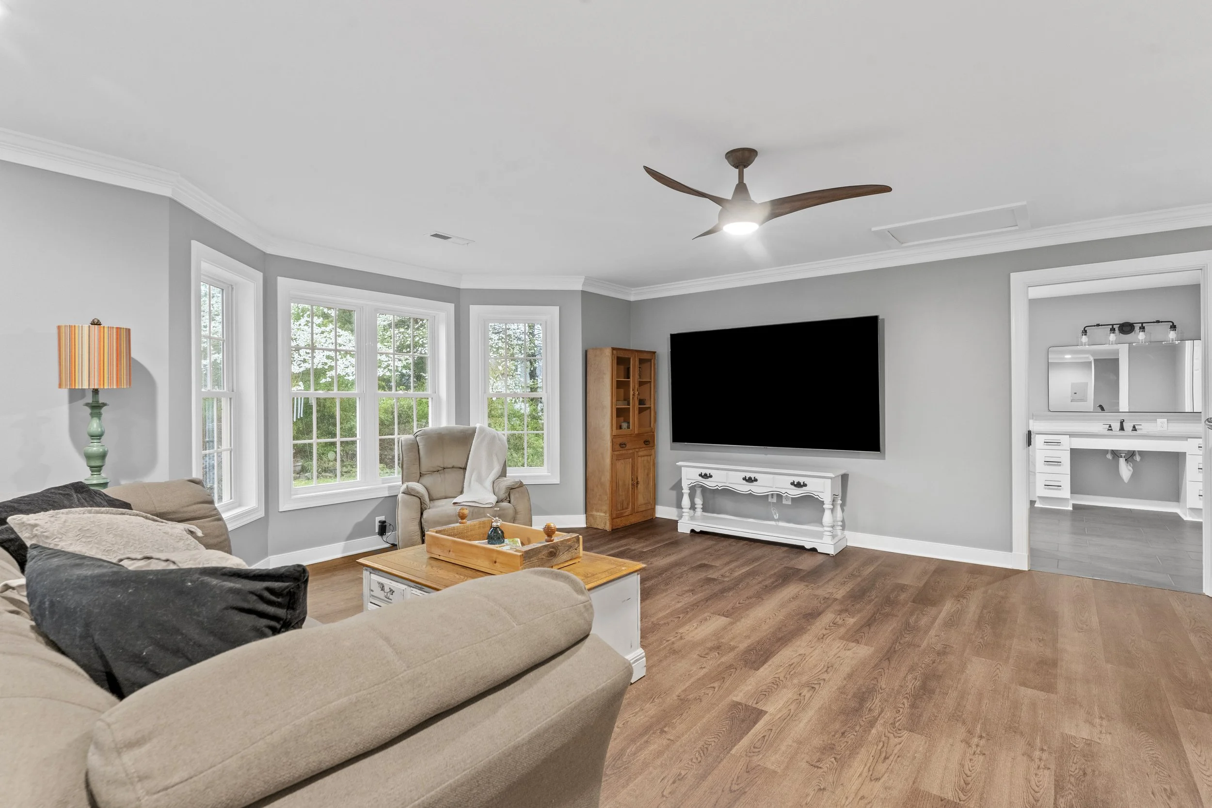 Living room with beige couch, armchair, large windows, flat-screen TV, and wooden accents.