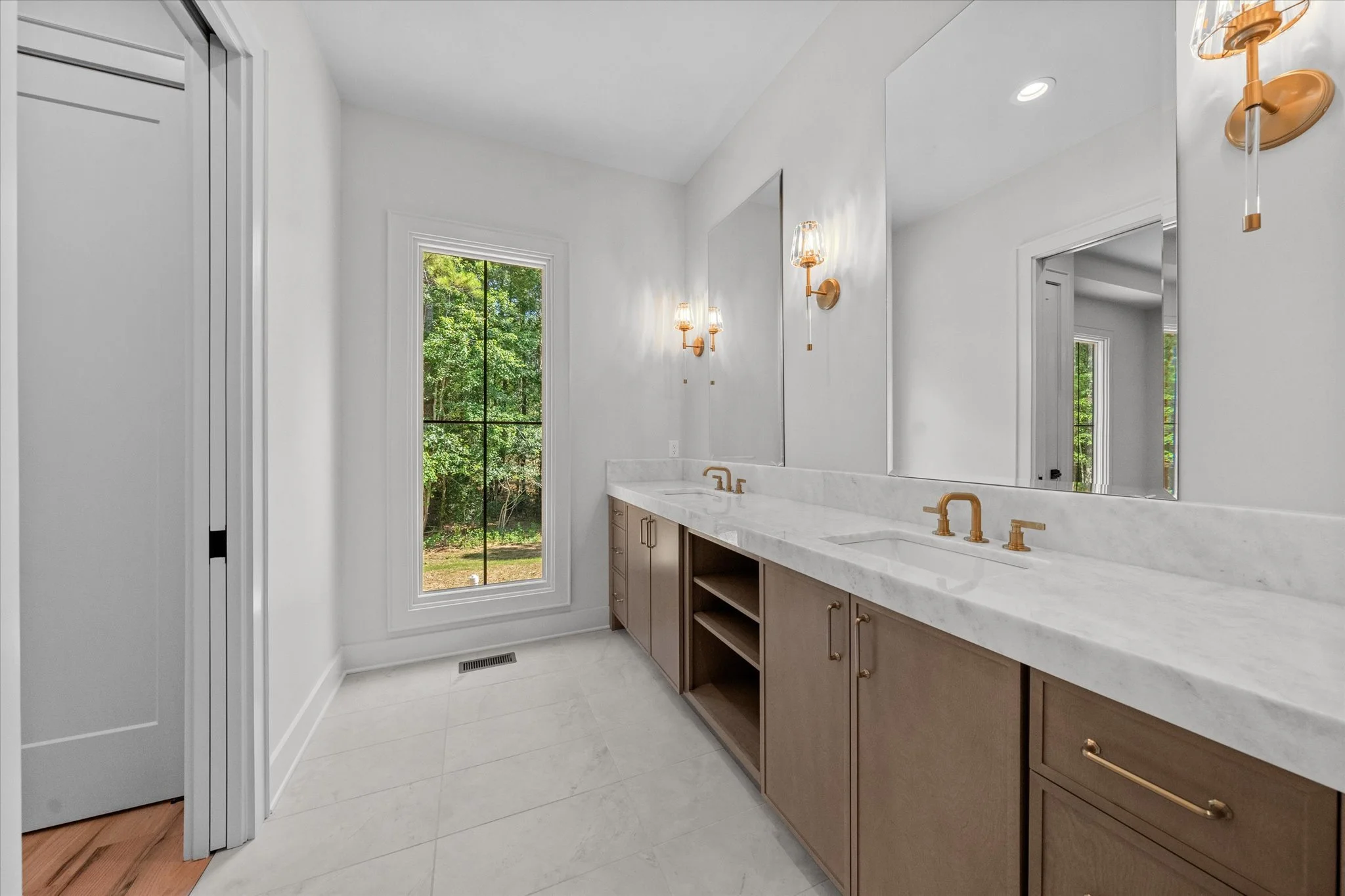 A modern bathroom with a double sink vanity, large mirror, gold fixtures, sconces, window showing green outdoor scenery, white walls, marble countertop, and light-colored wooden cabinets.