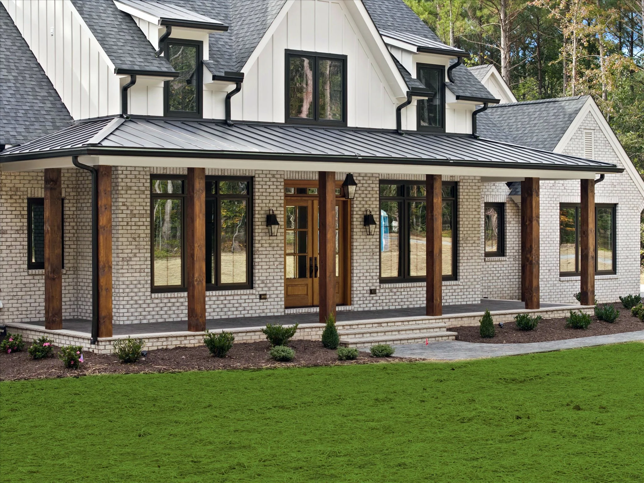 Front view of a modern two-story house with brick exterior, large windows, wooden columns, and a porch with steps, surrounded by a lawn and trees in the background.