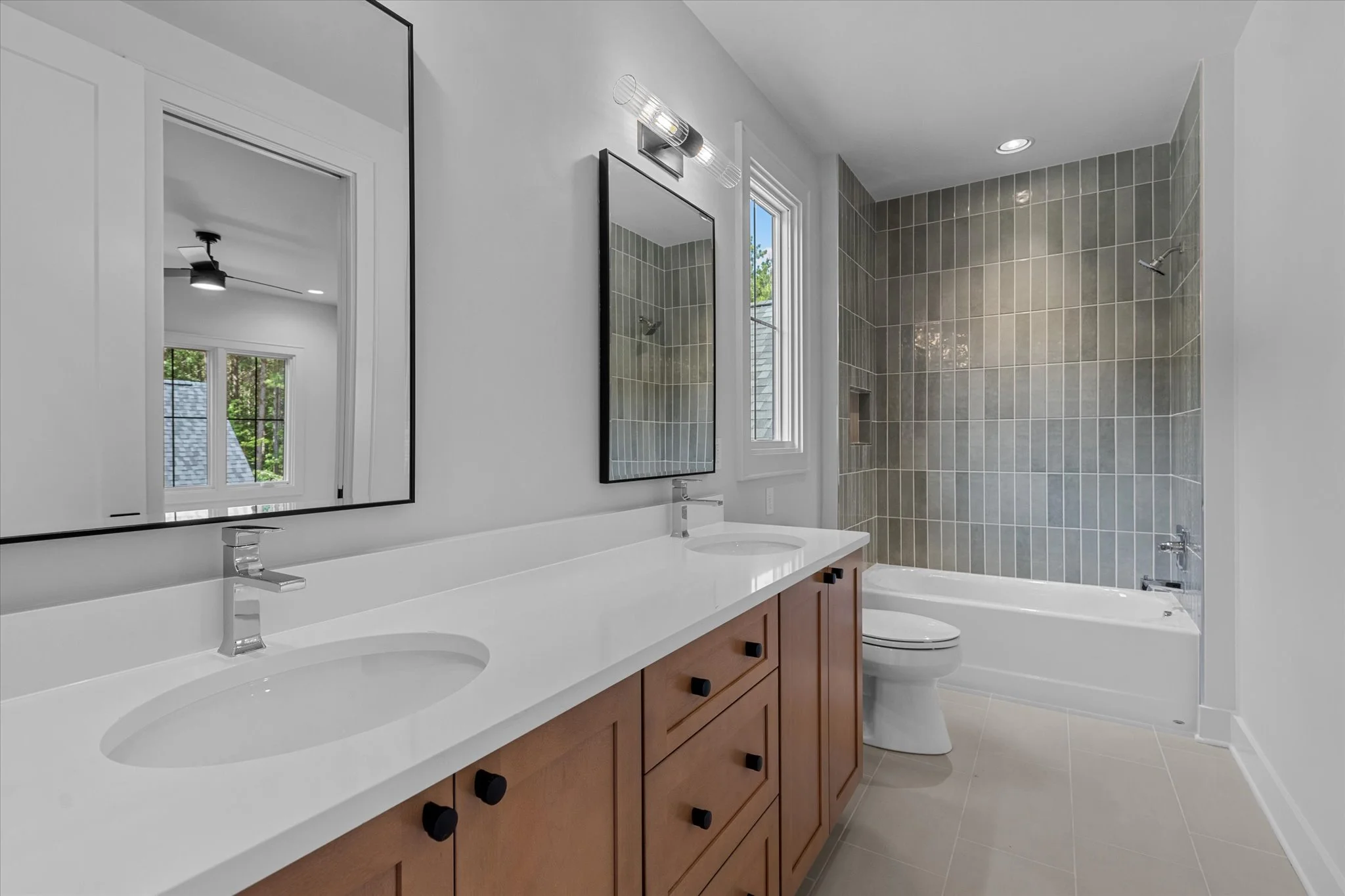 Modern bathroom with dual sinks, wooden vanity, large mirror, and a bathtub with gray tiled wall in the shower area, white walls, and light-colored floor tiles.