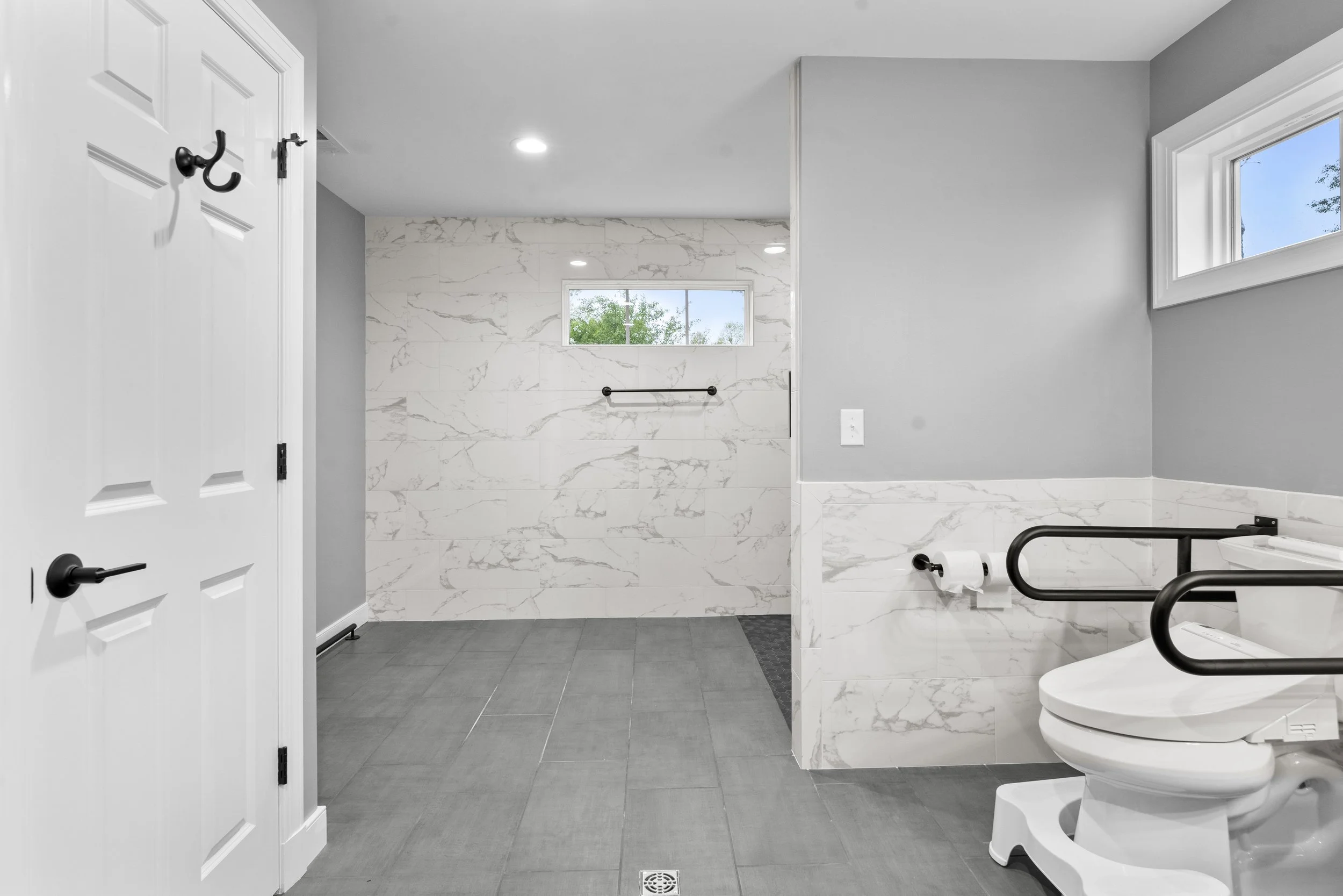 Modern bathroom featuring a toilet with black safety rails, gray tiled floor, white marble wall tiles, a small window, and a shower area with a window and black towel bar.