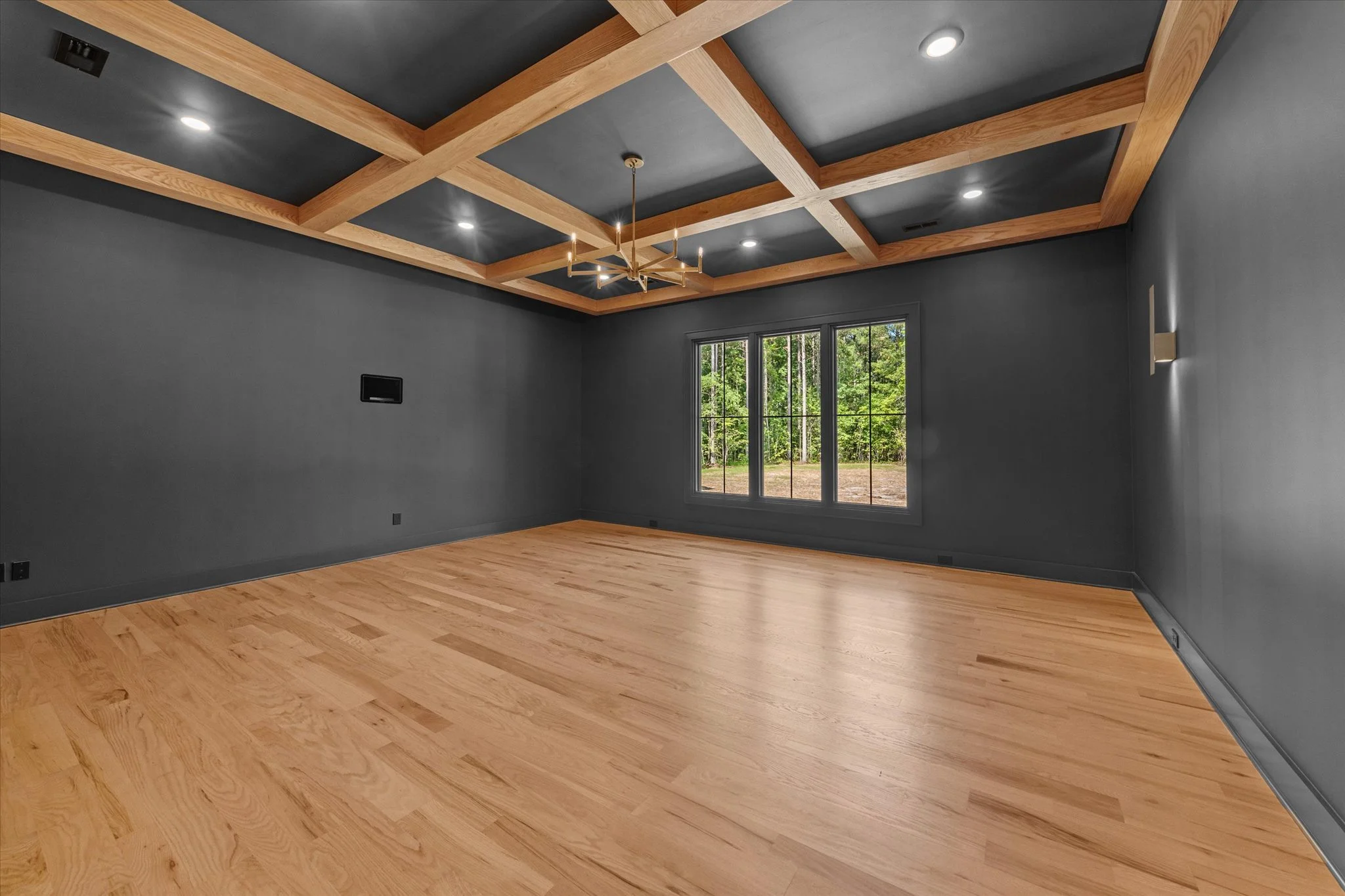 Empty room with dark gray walls, light-colored hardwood floors, a large window showing trees outside, and a ceiling with gray panels and a wooden grid pattern, featuring recessed lighting and a gold chandelier.