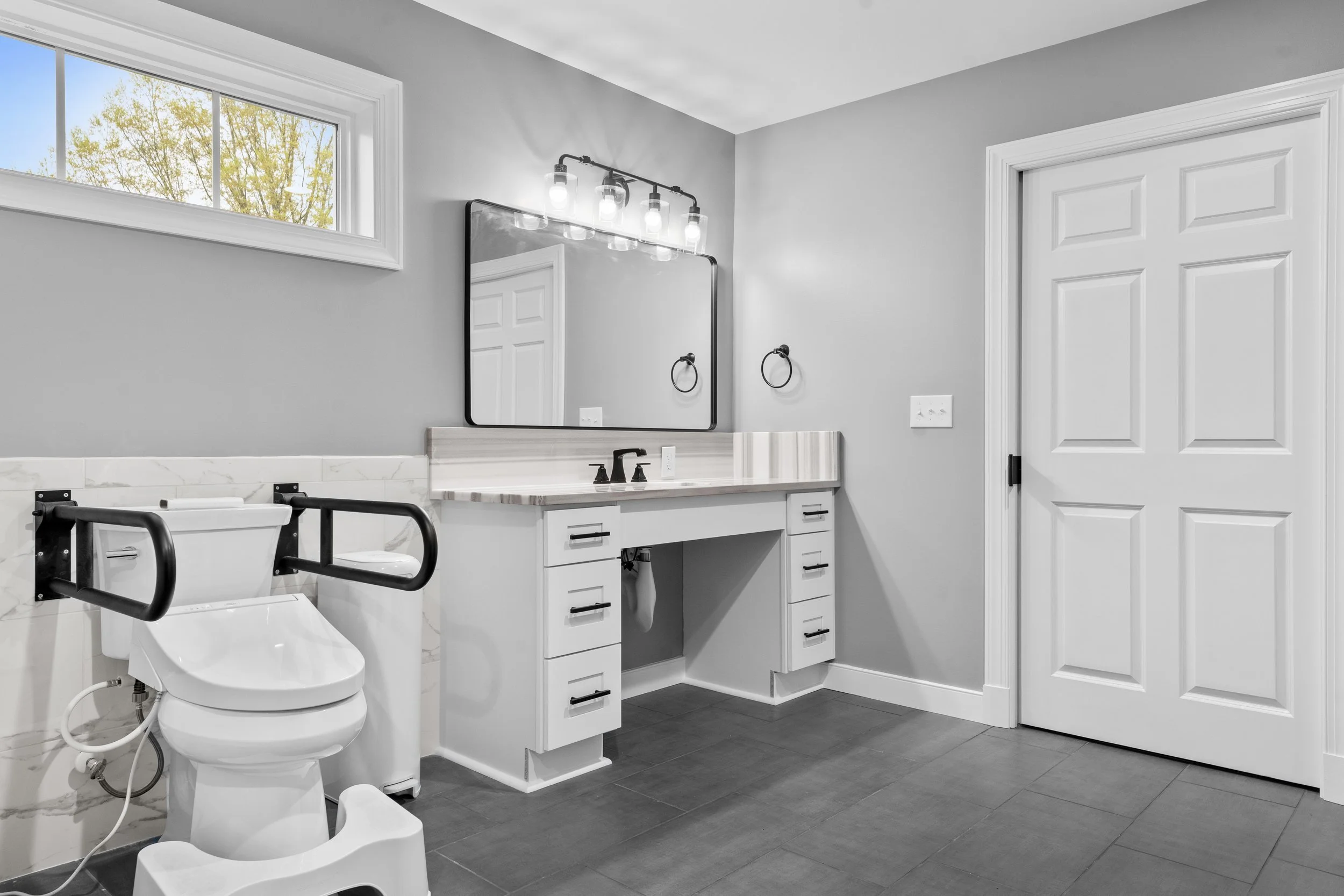 A modern, accessible bathroom with a toilet equipped with safety bars, a large window, a vanity with a mirror, and a closed white door.