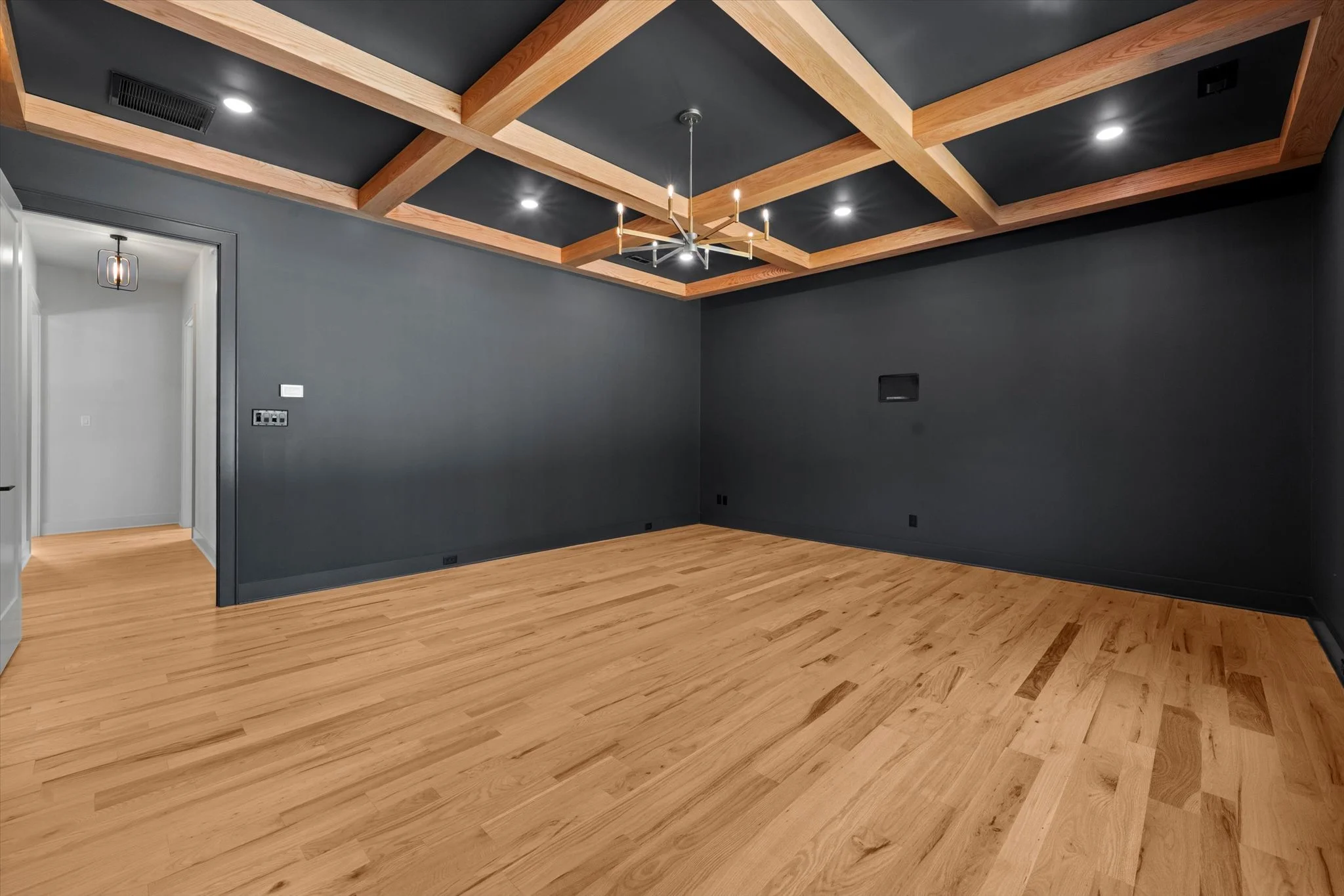 Empty room with dark gray painted walls, hardwood floors, and a ceiling with wood beams and a modern chandelier with candles.