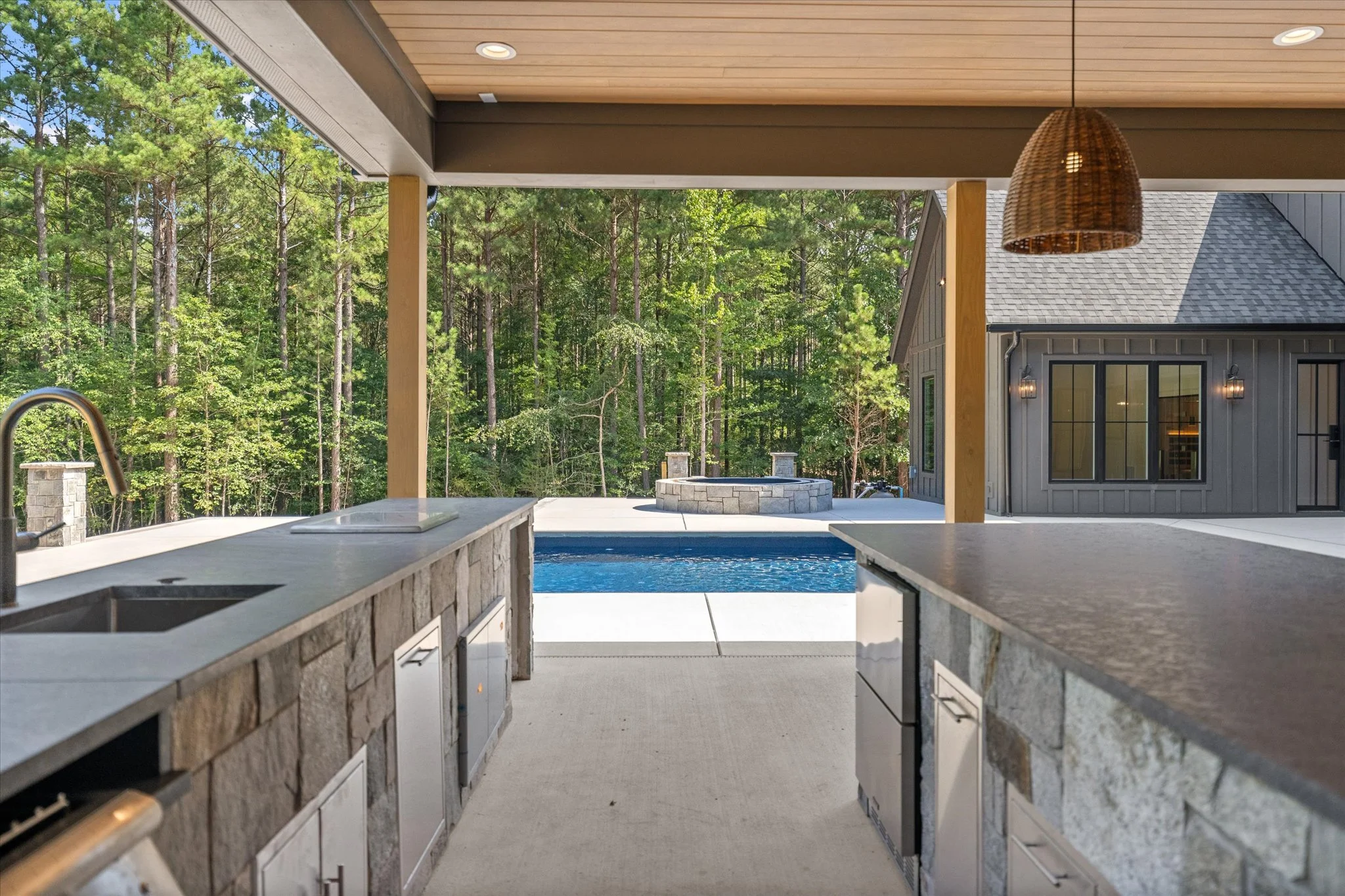 Outdoor kitchen with stone countertops, overlooking a swimming pool, with a wooded forest background and a fire pit in the distance.