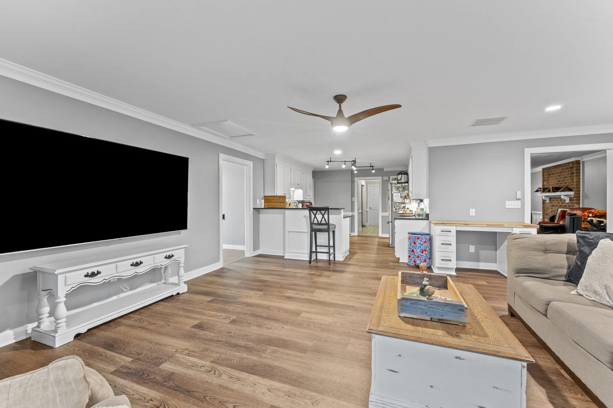 Open-concept living room with gray walls, hardwood floors, a large flat-screen TV, beige sofas, a wooden coffee table, a ceiling fan, and a kitchen area in the background.