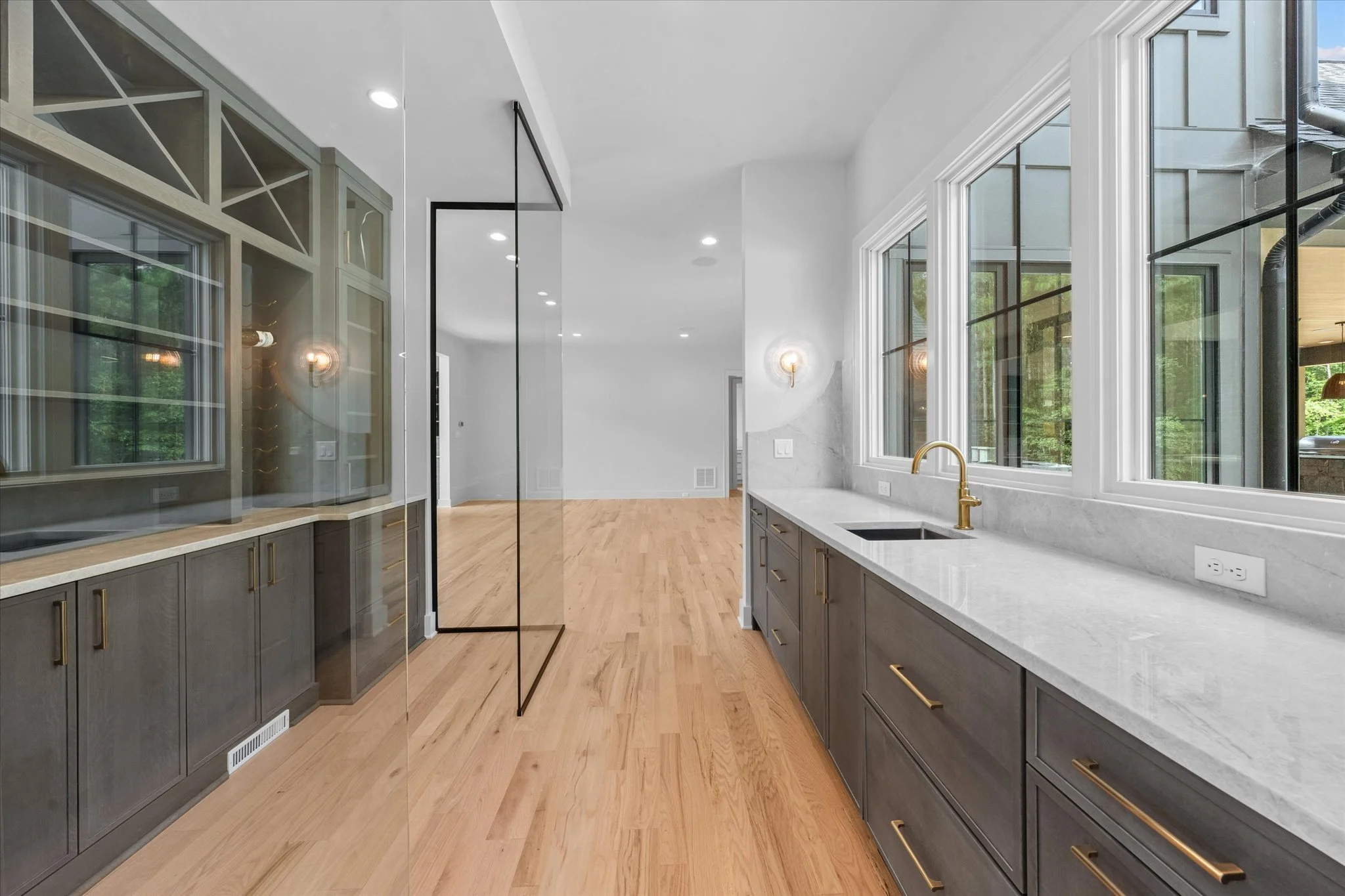 Modern kitchen with gray cabinets, white marble countertop, gold hardware, and large windows overlooking greenery.