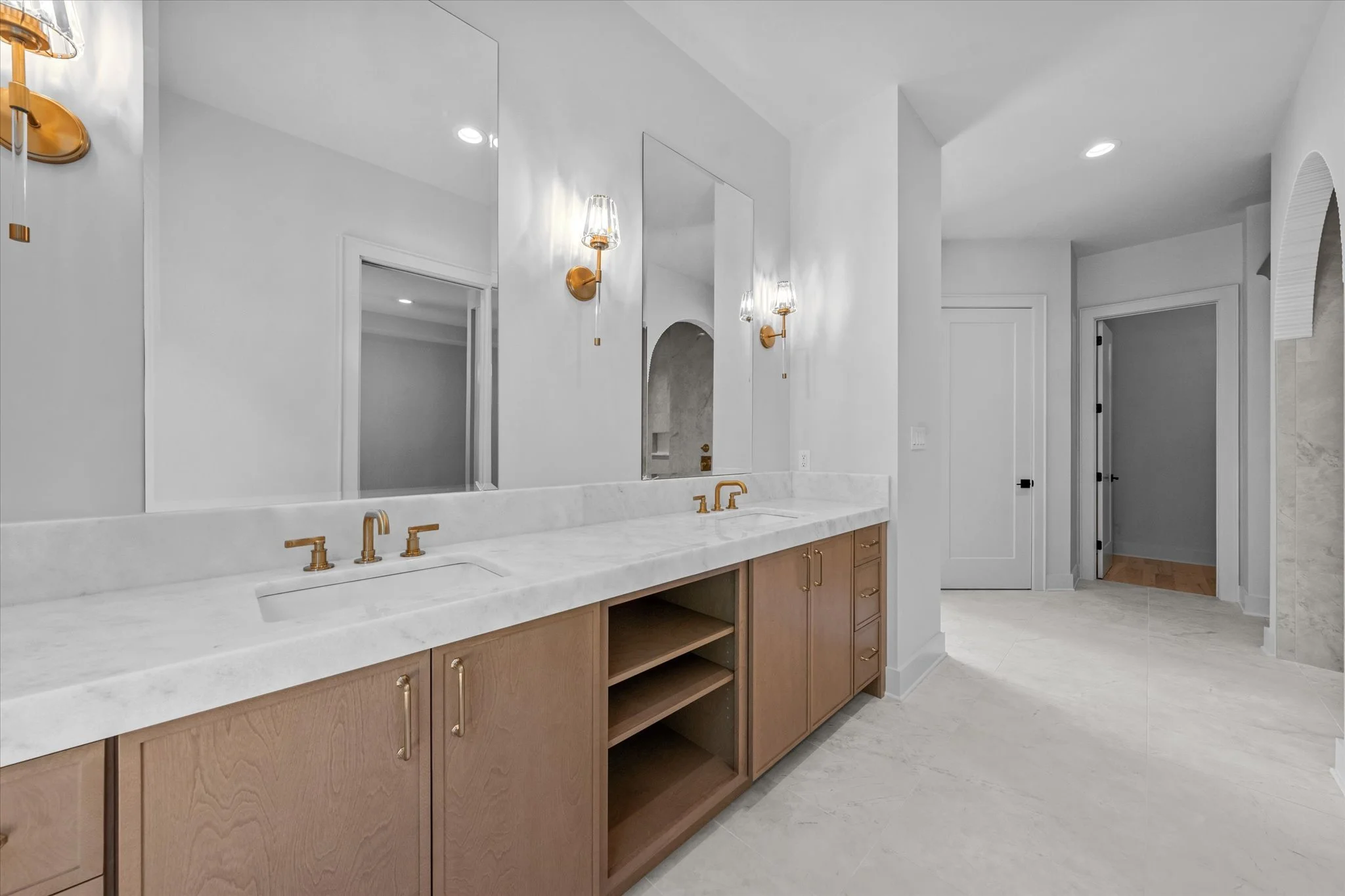 Modern bathroom with double vanity featuring a white marble countertop, gold fixtures, and wooden cabinets. Large mirror above the vanity, wall-mounted sconces, and light-colored flooring.