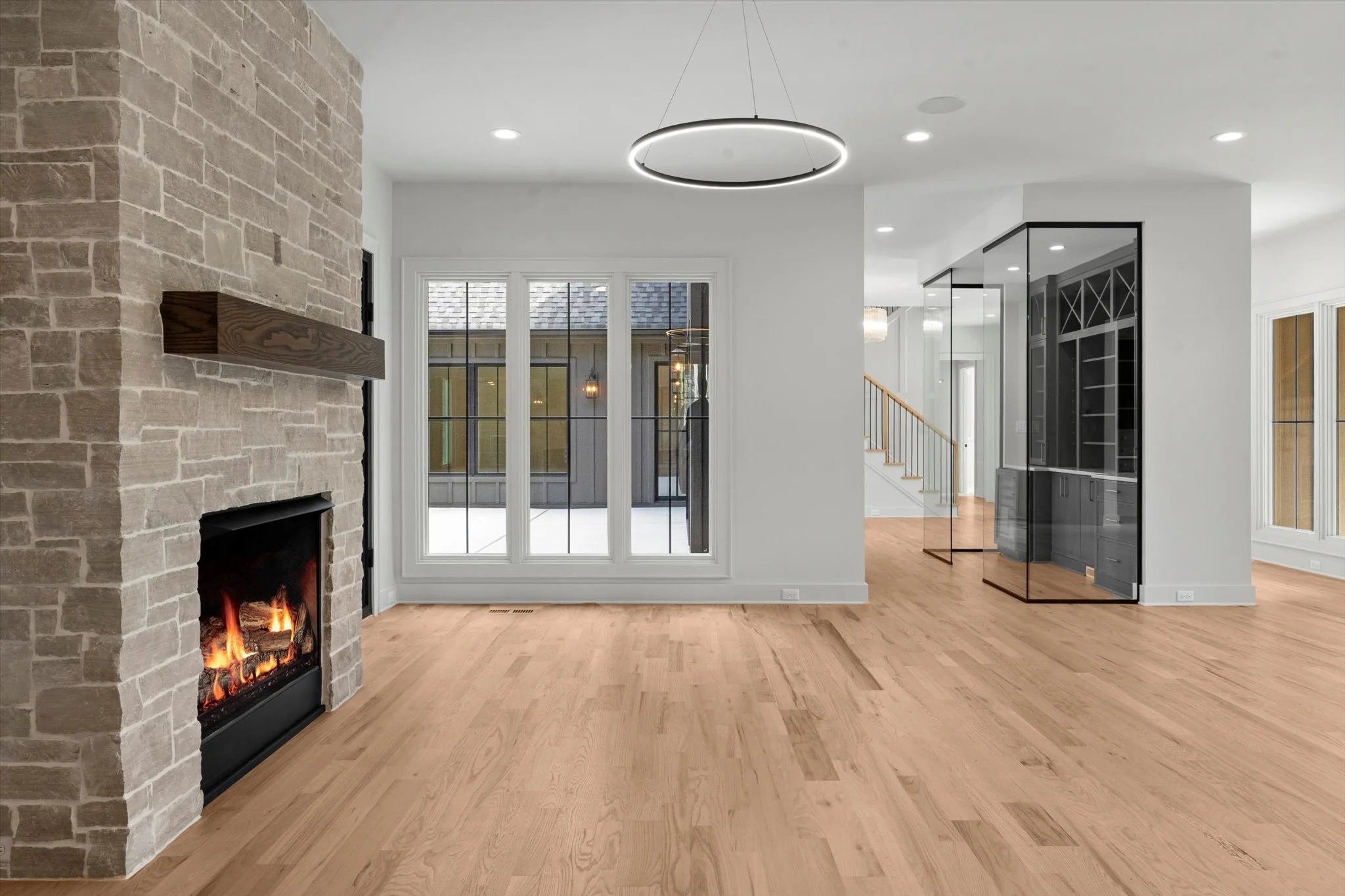 Empty living room with a stone fireplace, wood flooring, and large window looking into outdoor patio.