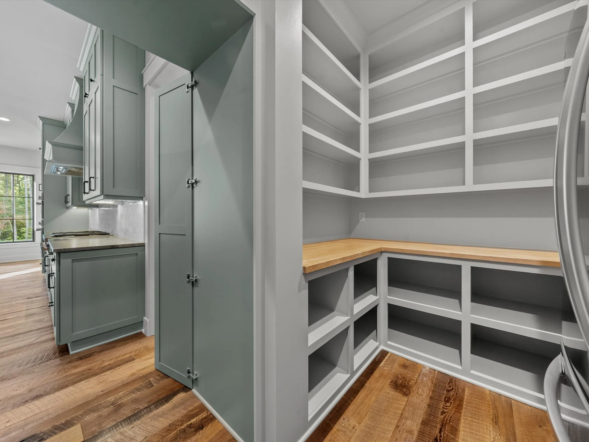 A kitchen with painted cabinets, a window, and a walk-in pantry with empty shelves and a wooden countertop.