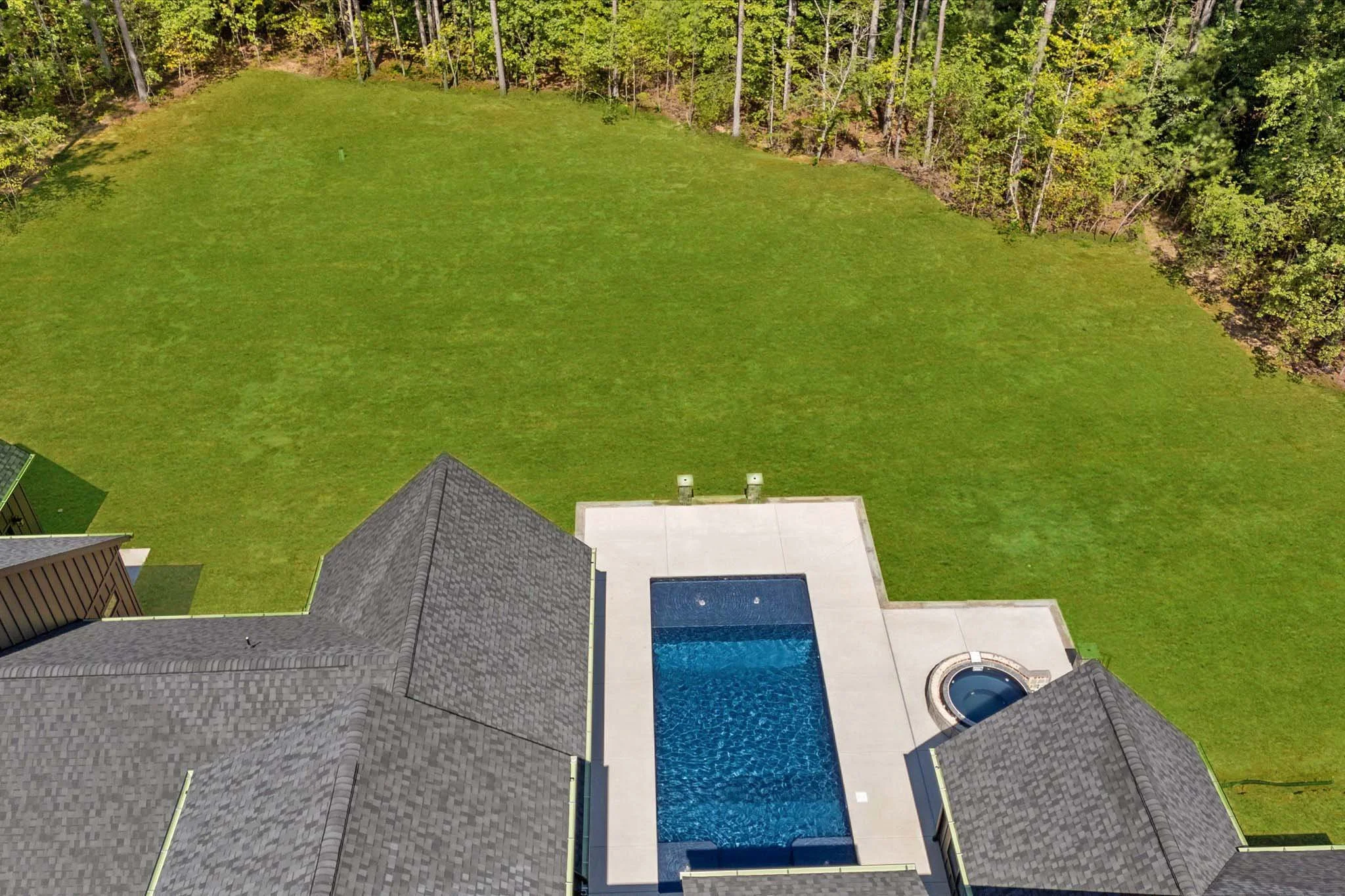 Aerial view of a backyard with a grassy lawn, a swimming pool, a hot tub, and surrounding trees.