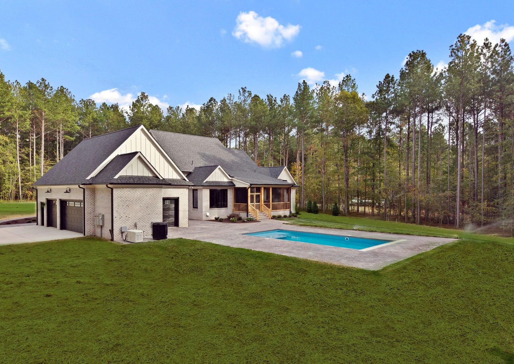 A modern house with a dark gray roof, white brick exterior, and a wooden porch, situated in a grassy backyard with a swimming pool, bordered by tall trees under a blue sky with scattered clouds.