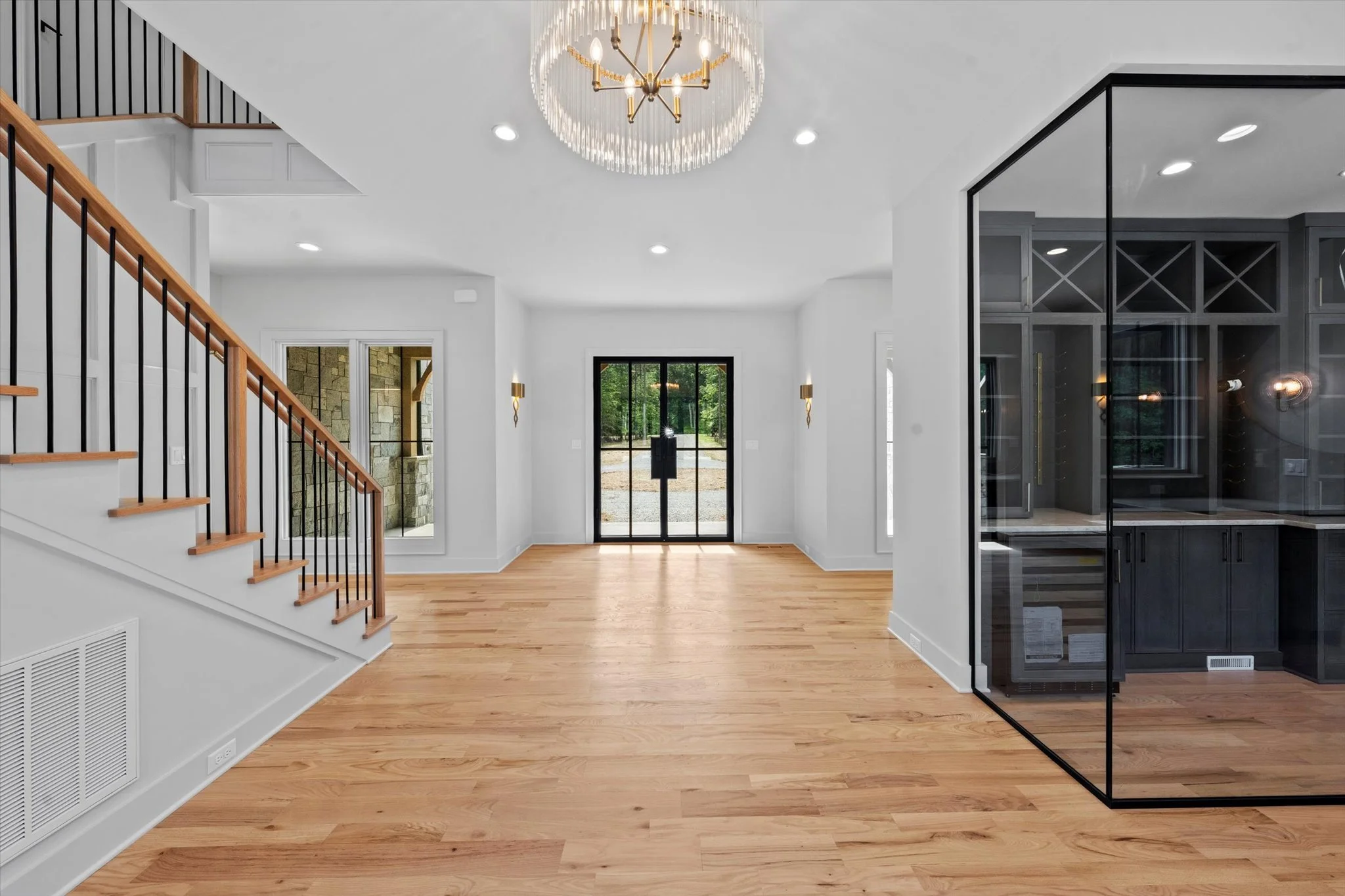 Modern living space with hardwood floors, white walls, black-framed glass front door, a chandelier, and a glass-walled office area on the right.