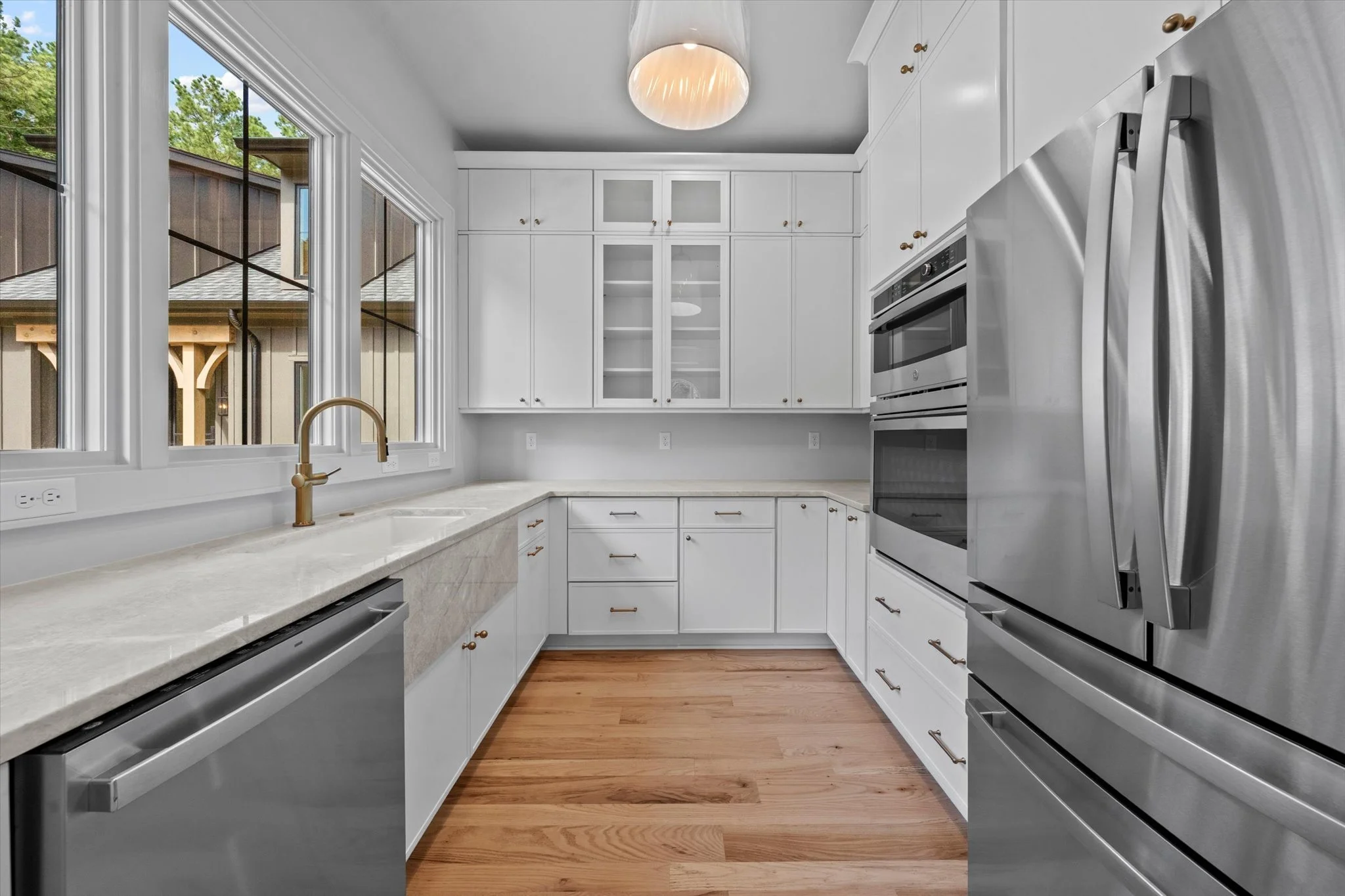 Modern kitchen with white cabinets, a marble countertop, and stainless steel appliances near large windows with a view of a house and trees outside.