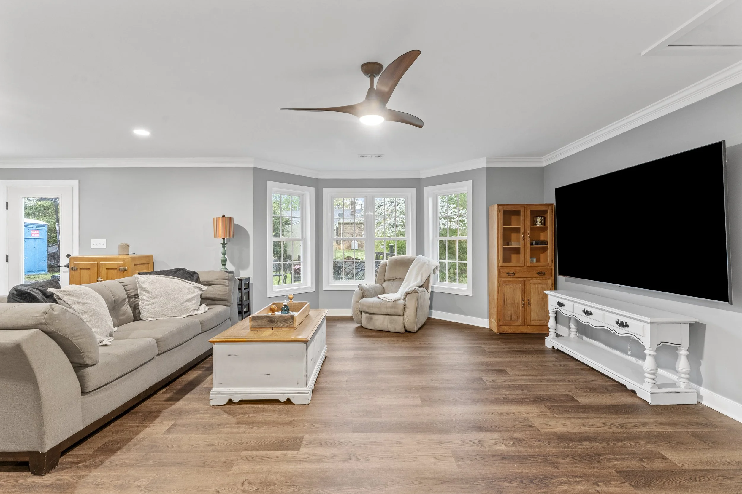 Living room with beige couch, armchair, wooden cabinets, large flat-screen TV, white coffee table, and hardwood flooring with gray walls and white trim.