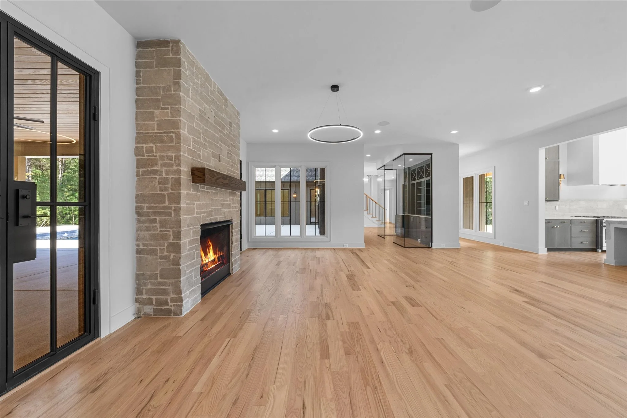 Empty living room with hardwood floors, a stone fireplace with a wooden mantel, and large windows with a view of the backyard. Modern lighting fixtures and a glass cabinet are also visible, with an open kitchen area on the right.