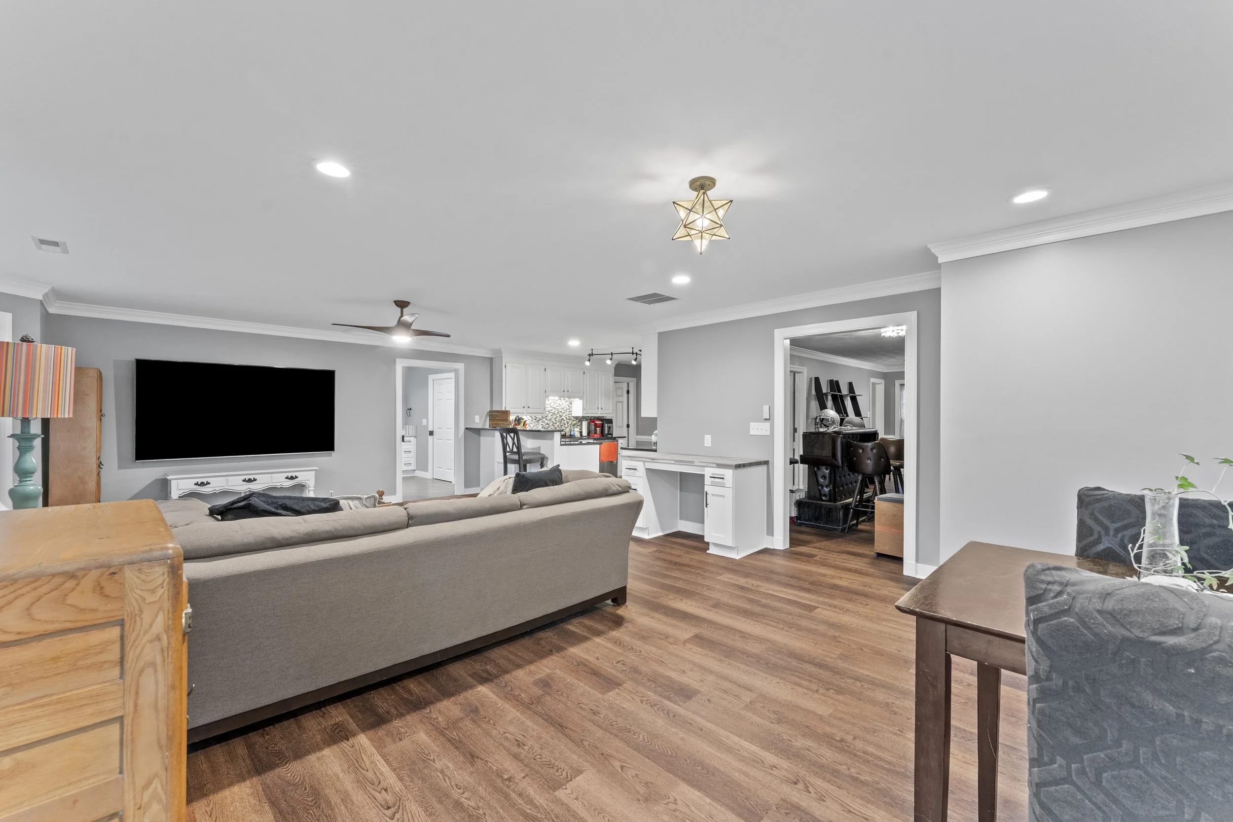 Open-concept living room with gray walls, hardwood floors, a large beige sofa facing a wall-mounted flat-screen TV, a side table with a lamp, a small bar area with chairs, and a kitchen in the background with white cabinets and a mosaic tile backspla