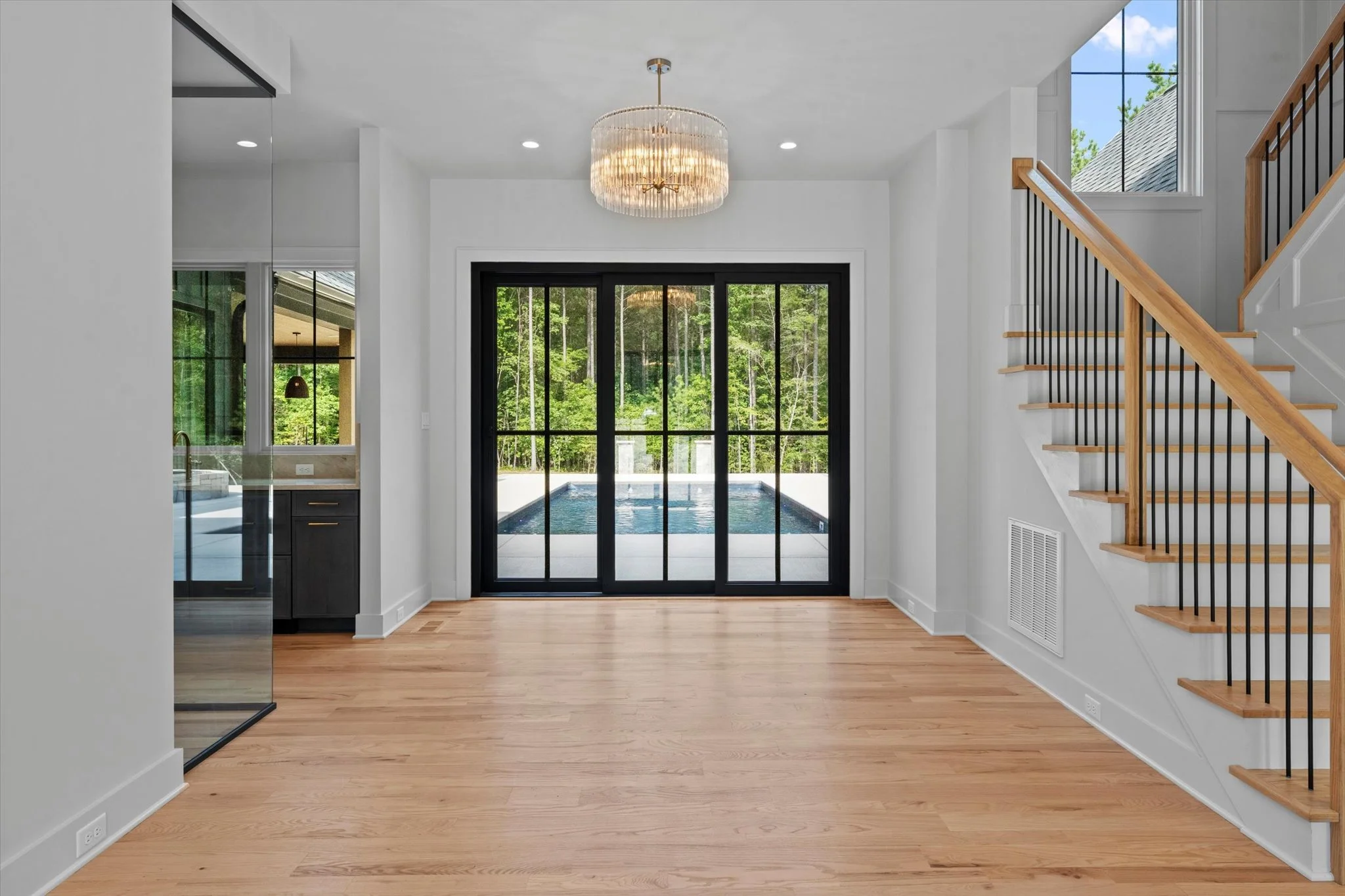 Empty living space with hardwood floors, white walls, a round chandelier, black-framed sliding glass doors leading to a backyard with a pool, and a staircase with wooden handrails and black balusters.